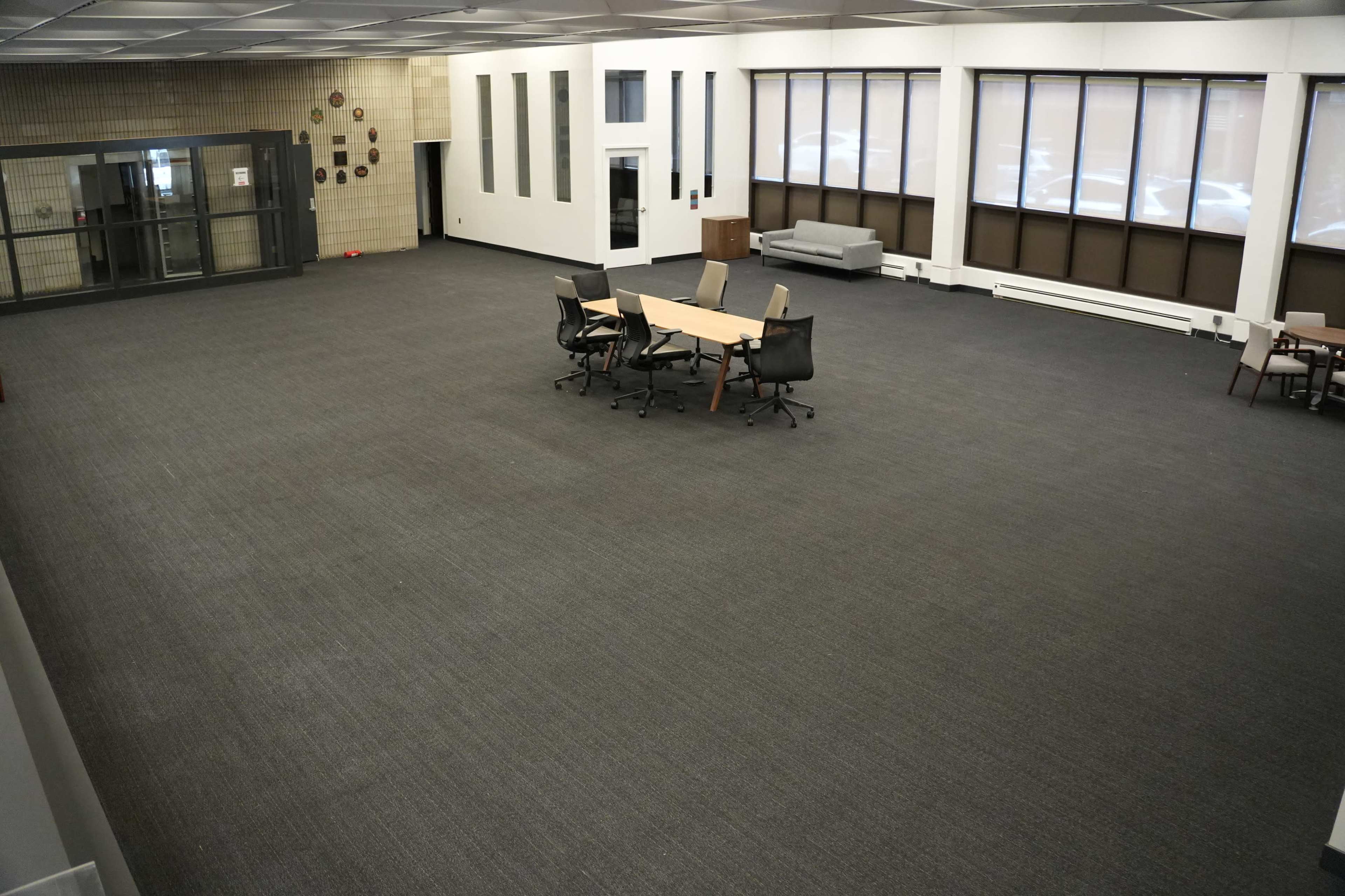 An empty, well-lit room features several tables and chairs arranged in the center, with large windows along one wall and an elevator to one side.