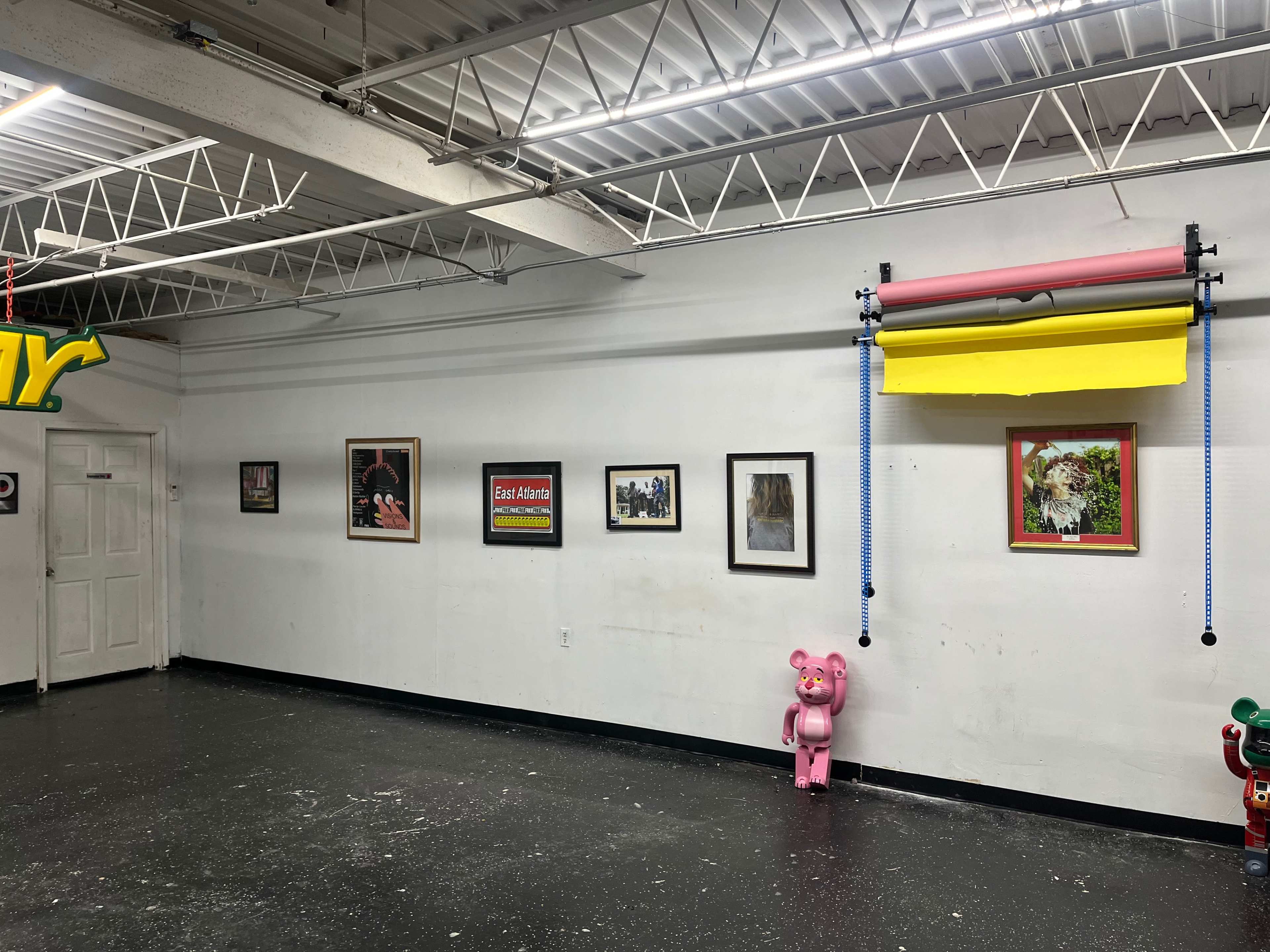 The image shows a gallery-like space with framed artworks and photographs displayed on a white wall, accompanied by a pink bear sculpture and multicolored window shades.