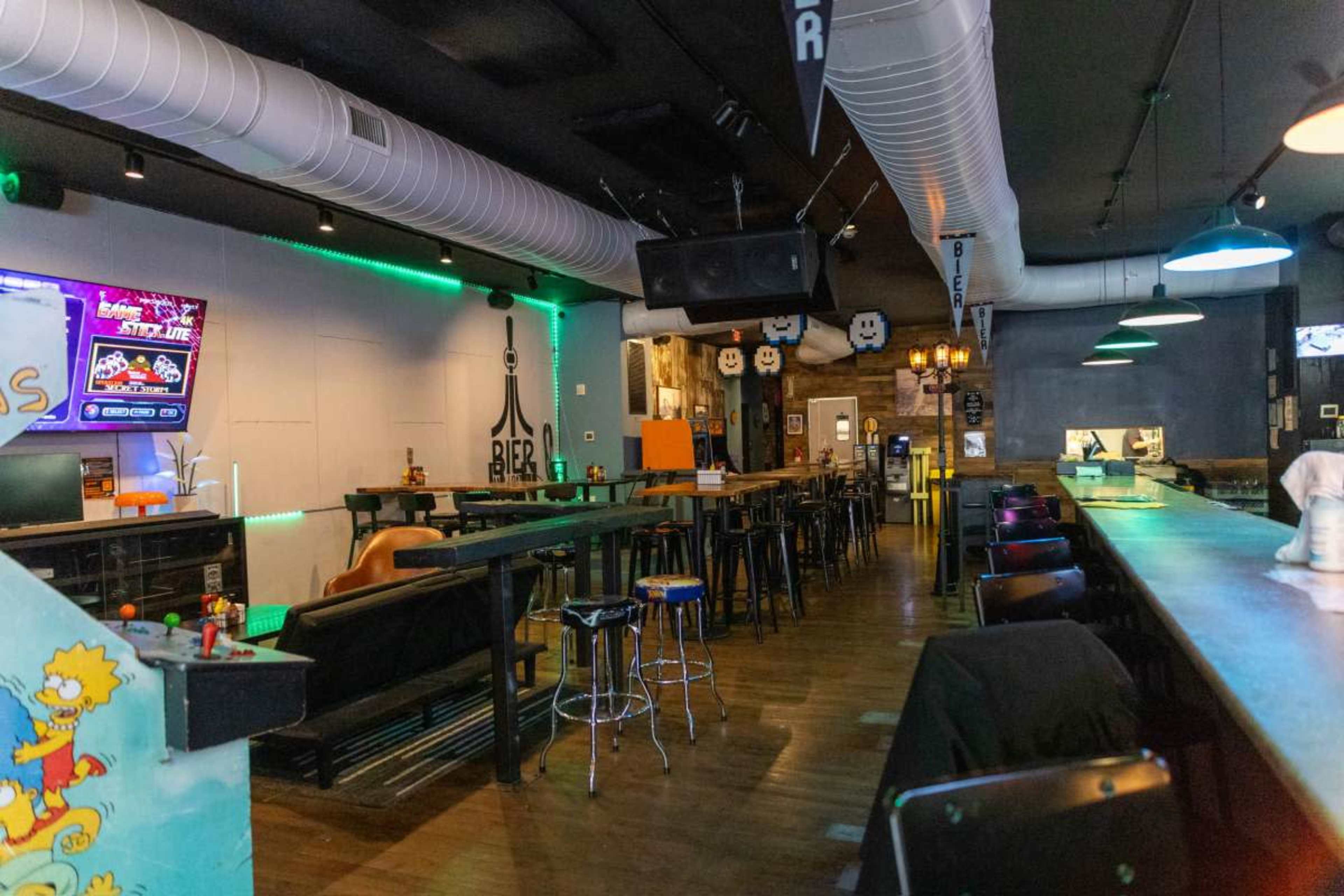 The image shows the interior of a bar with high tables, stools, and a long counter, featuring neon lights and a bar game area.