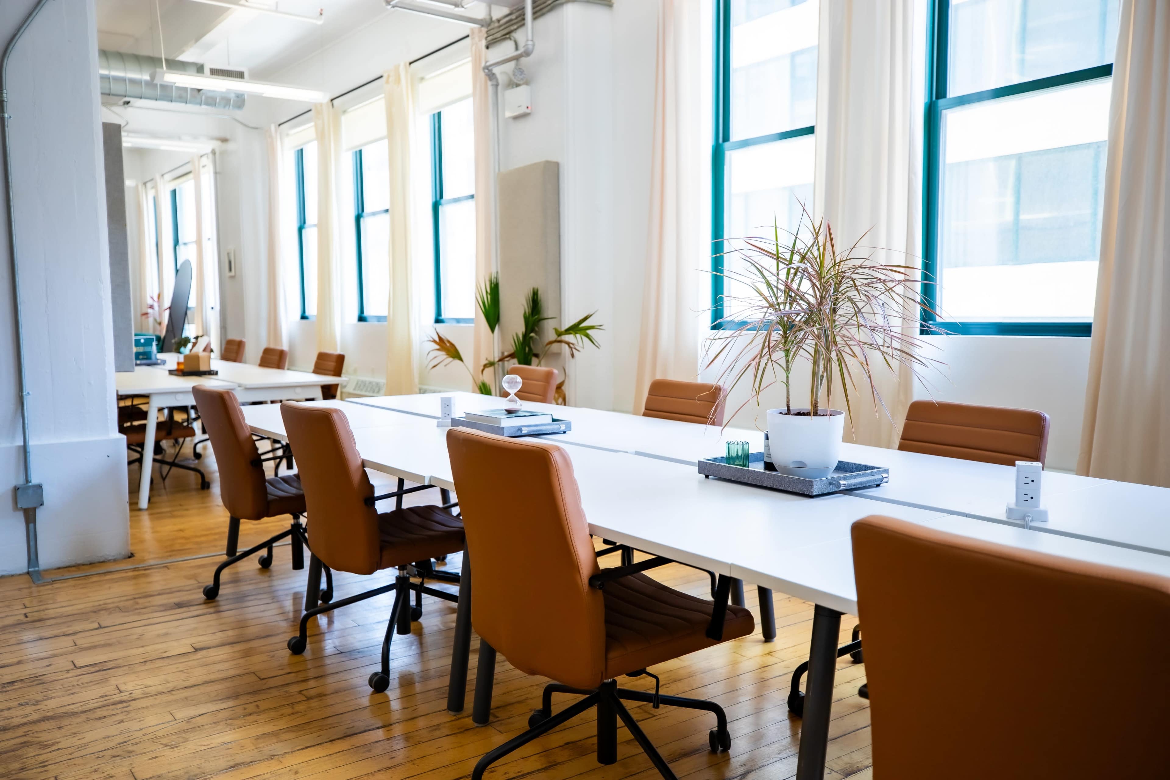 A spacious office features a long table surrounded by brown leather chairs, with large windows allowing natural light to illuminate the room.