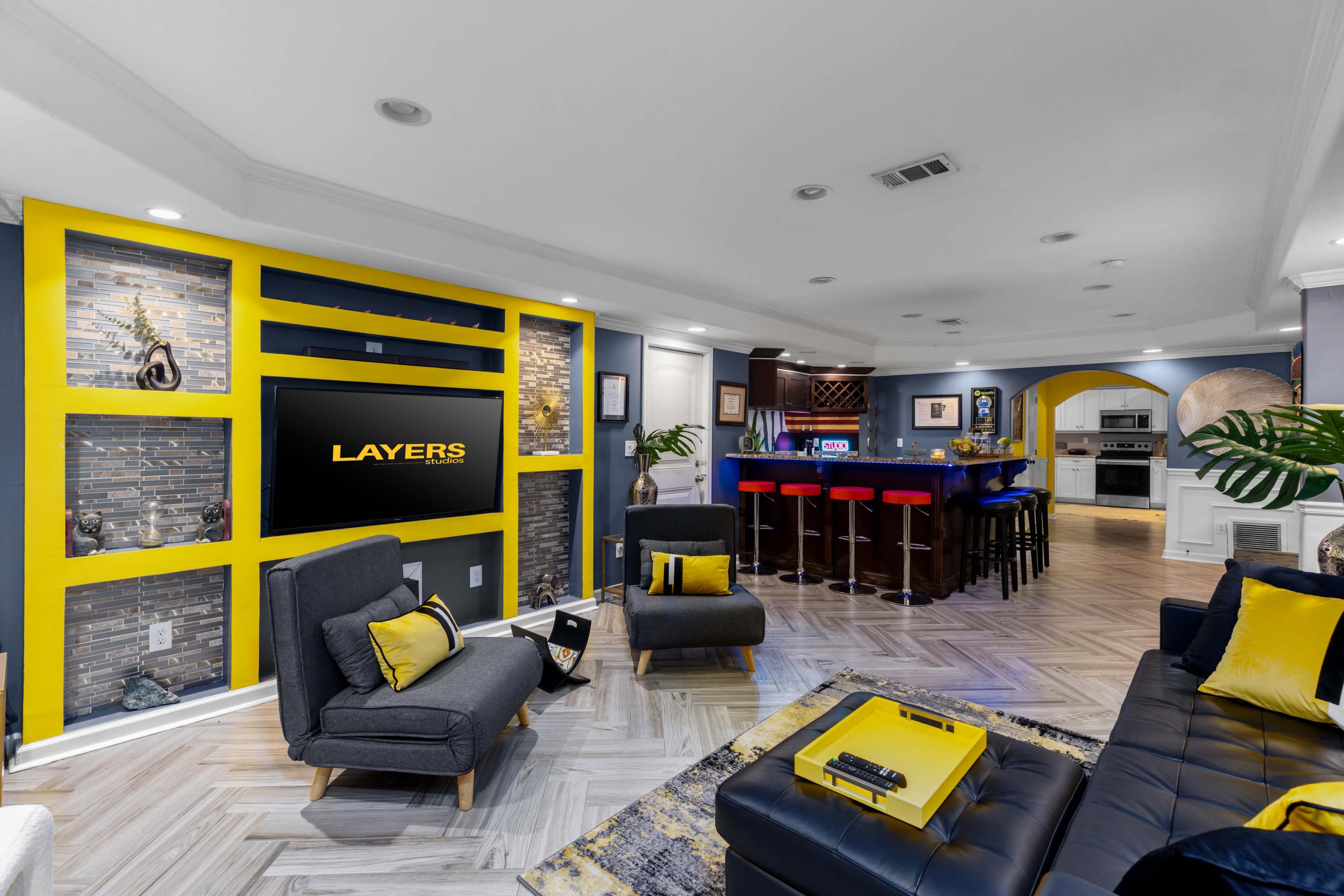 The modern living room features a yellow accent wall with a mounted TV, contrasting with dark furniture and an adjacent kitchen area with a bar and seating.