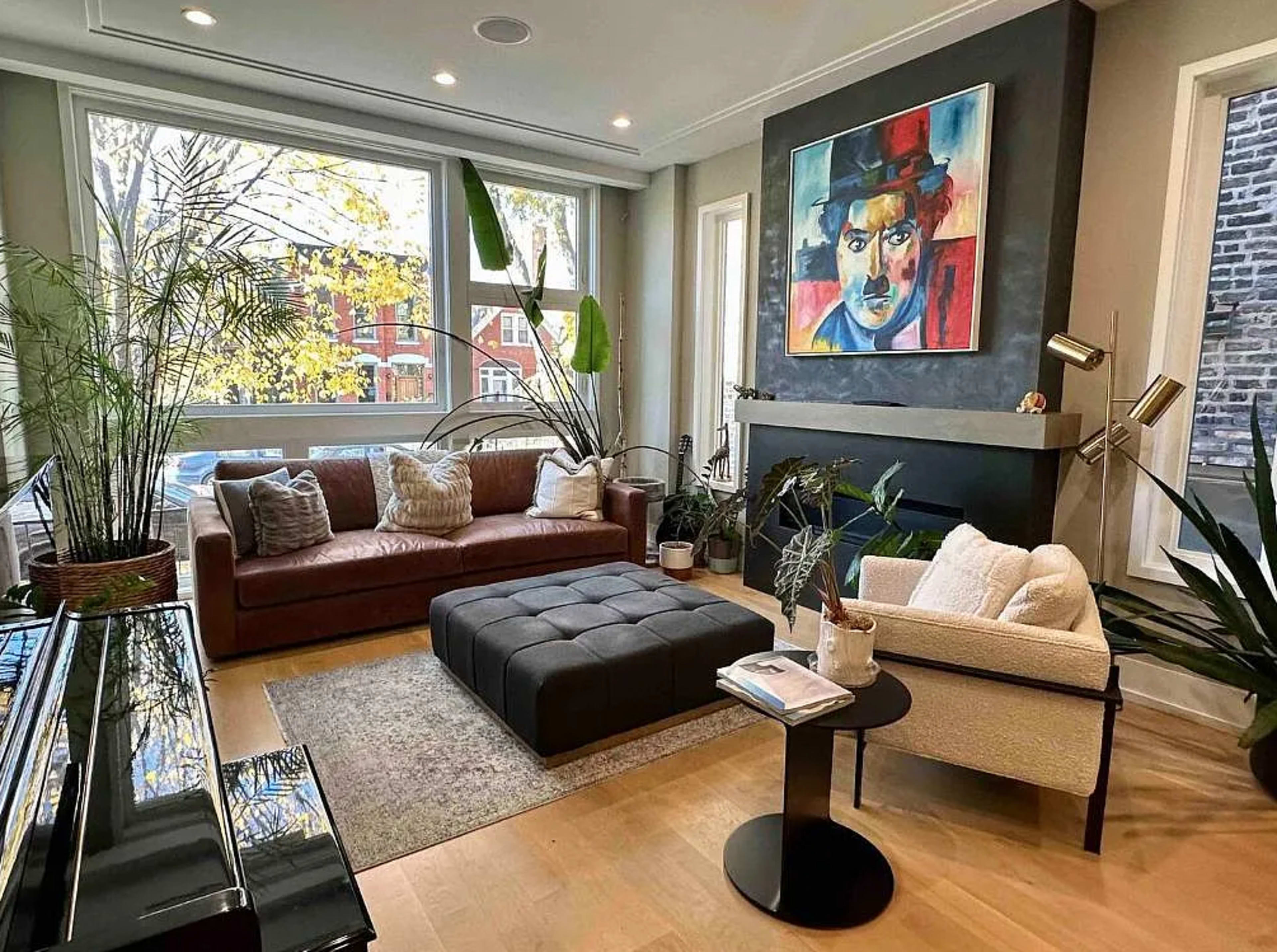 The image features a modern living room with a brown leather sofa, a white armchair, a coffee table, and a large painting on the wall, surrounded by indoor plants and large windows.