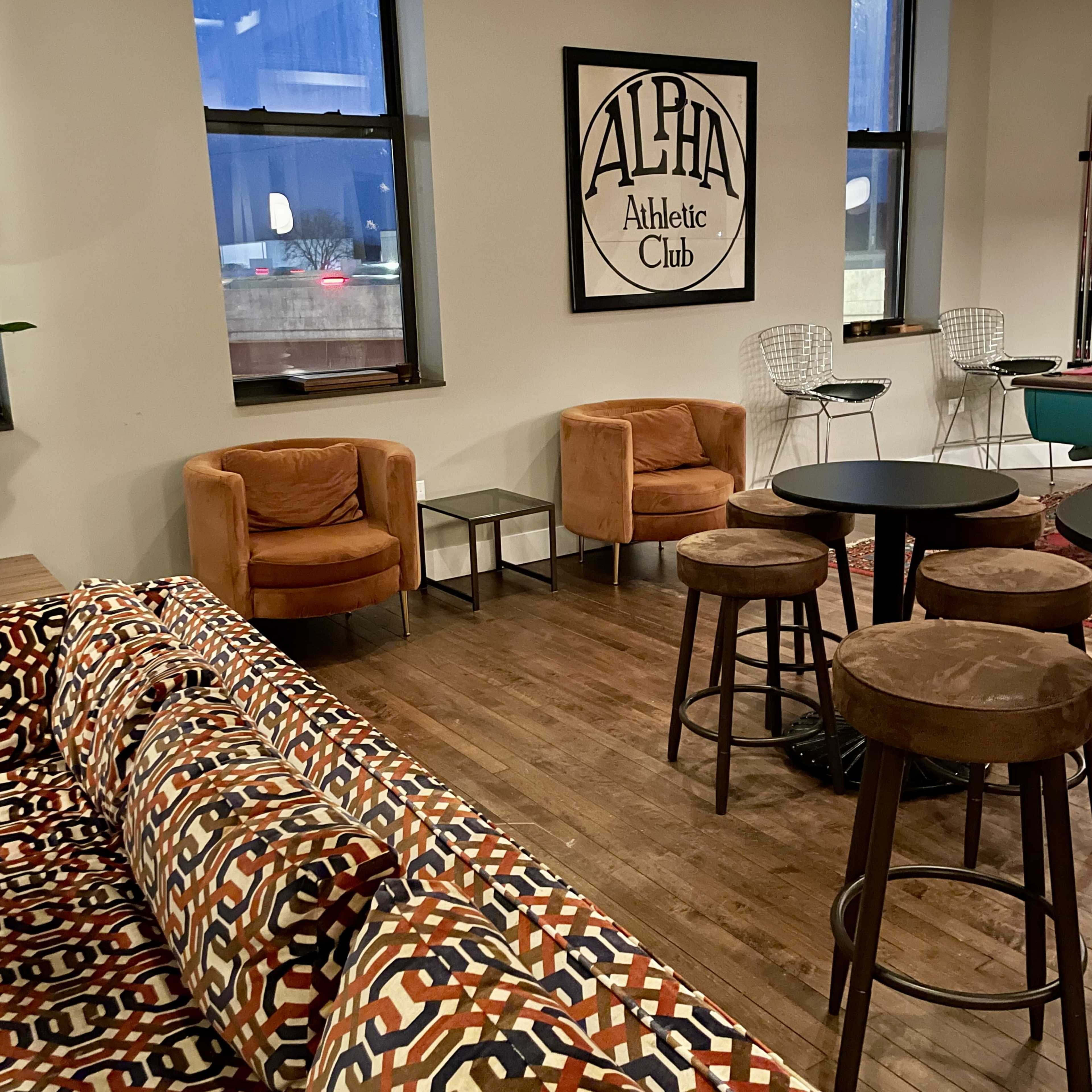 The image shows a cozy interior of a lounge area featuring two orange armchairs, a dark round table, bar stools, and a patterned sofa, with a wall art piece that reads "ALPHA Athletic Club."