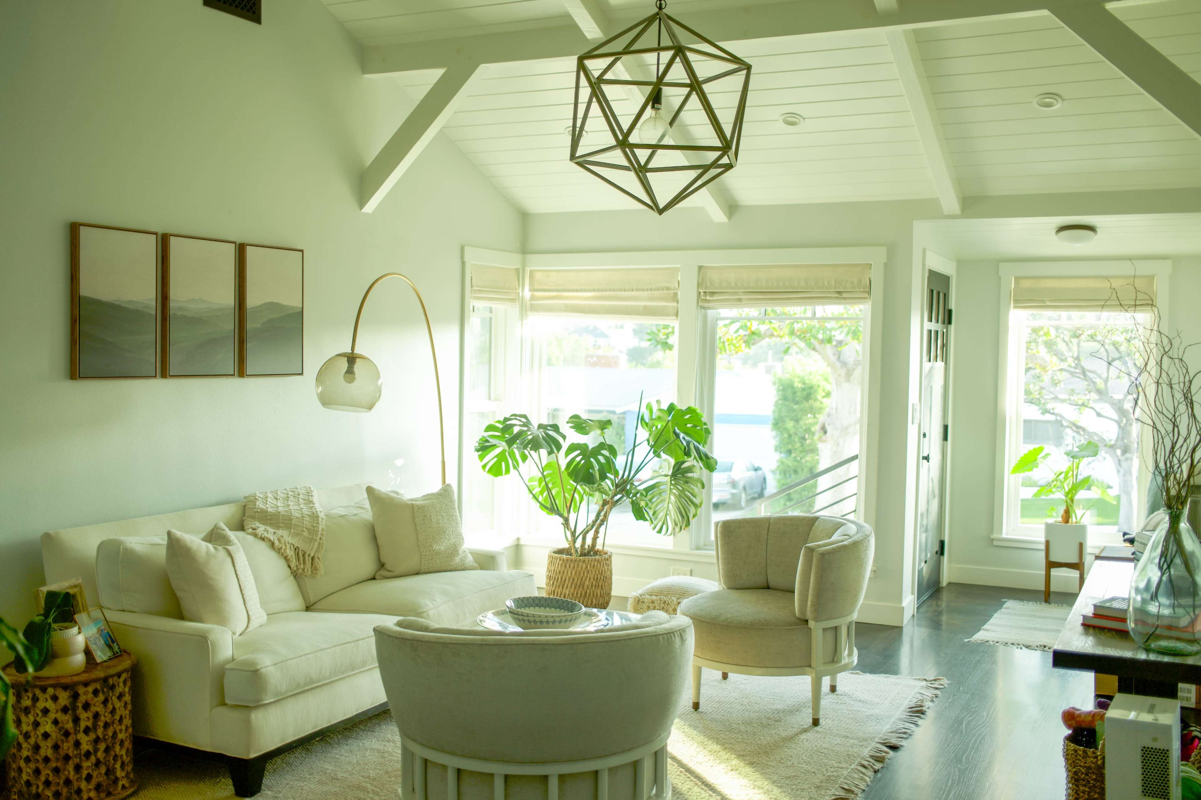 The image shows a light-filled living room featuring a white sofa, two armchairs, and a large potted plant under a geometric light fixture.