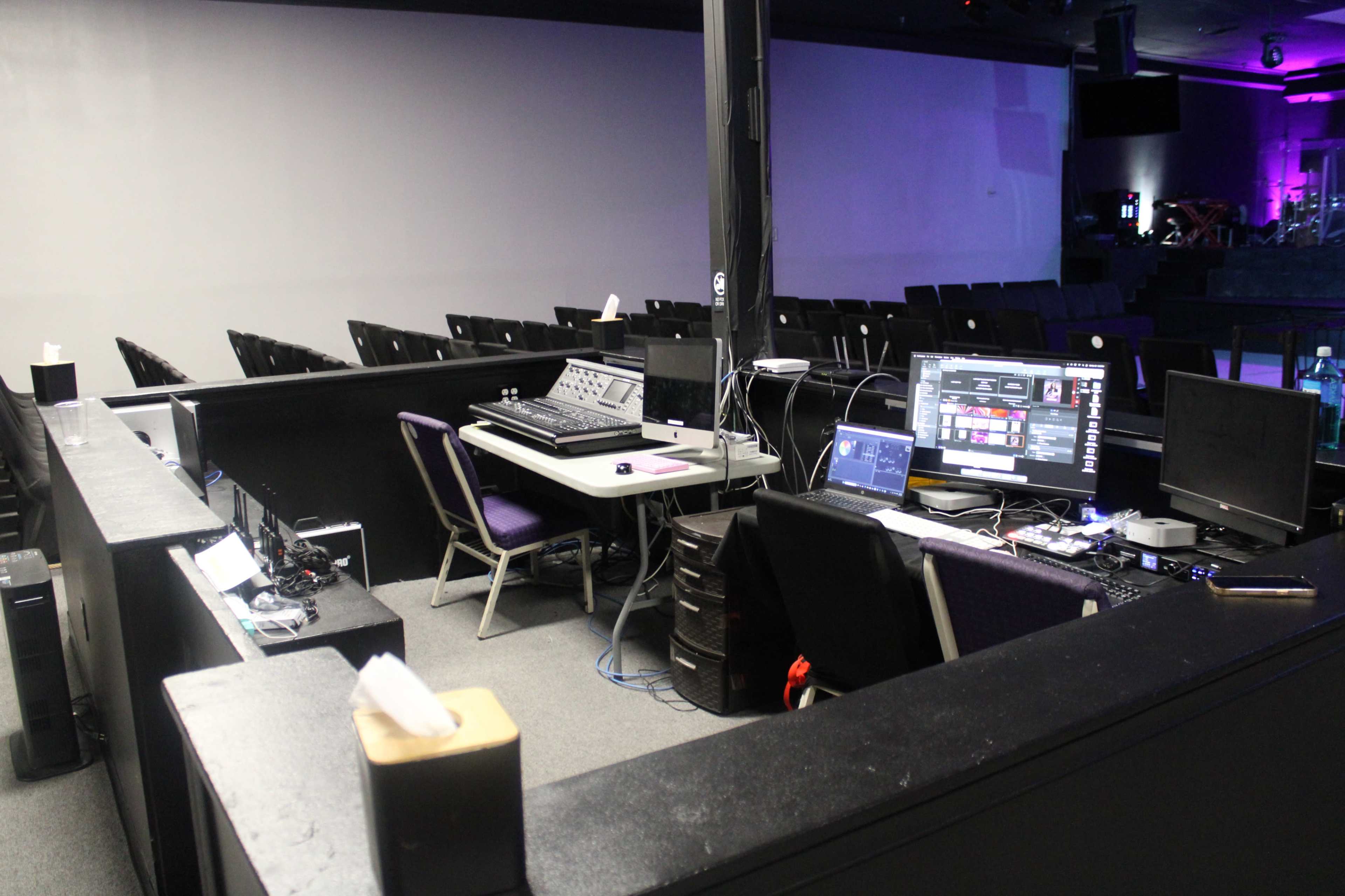 The image shows a sound and lighting control booth in a venue, equipped with multiple monitors, audio equipment, and chairs arranged in the background.