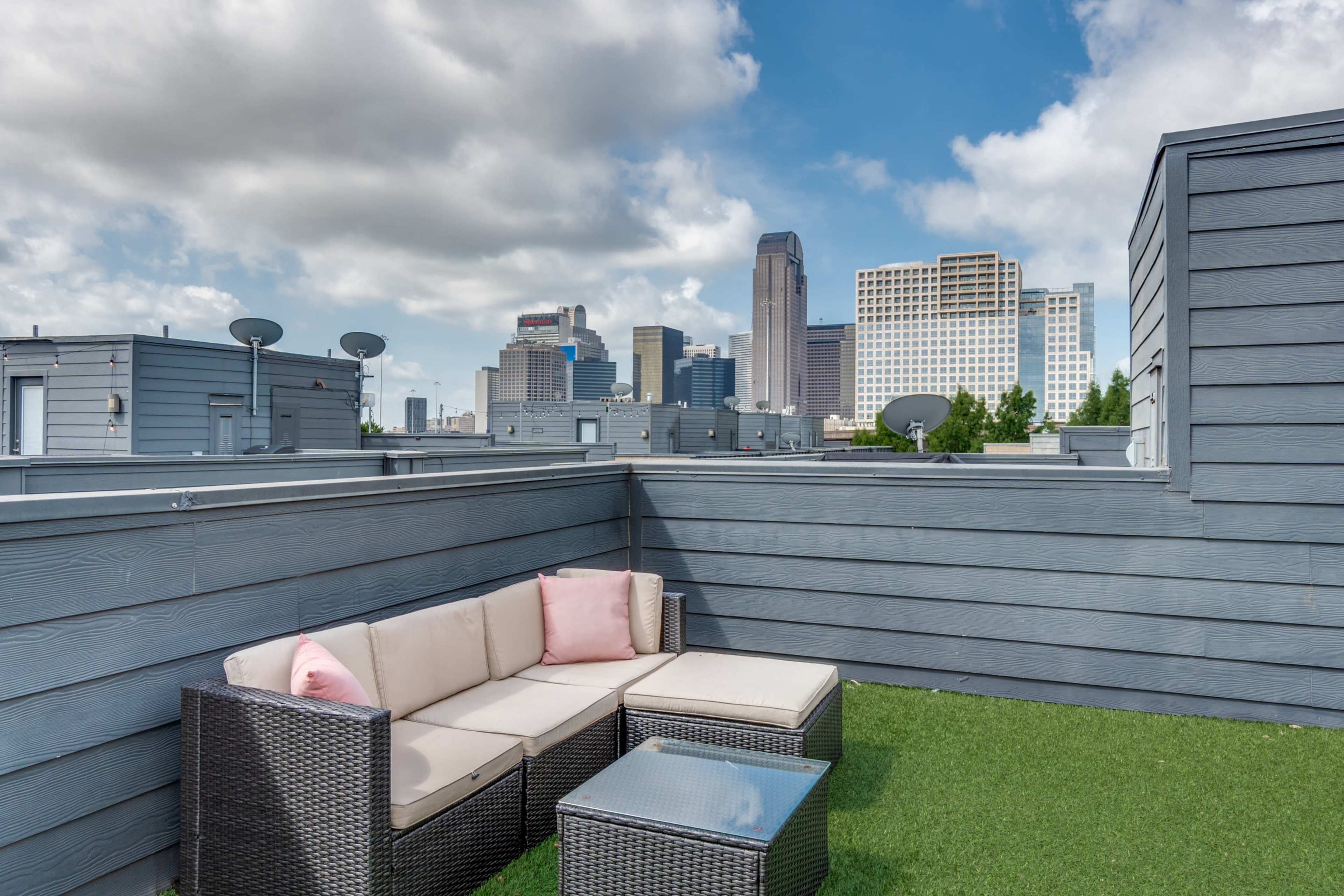 A rooftop lounge area with a sectional sofa and coffee table overlooks a city skyline under a partly cloudy sky.