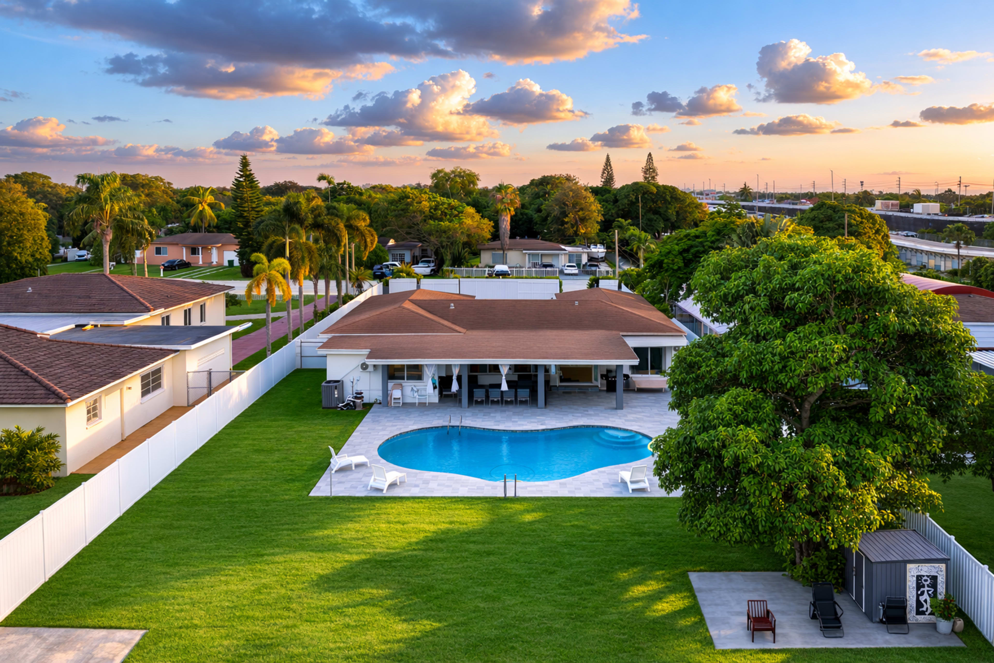 A well-manicured backyard features a swimming pool surrounded by grass, with several palm trees and homes visible in the background under a colorful sunset sky.