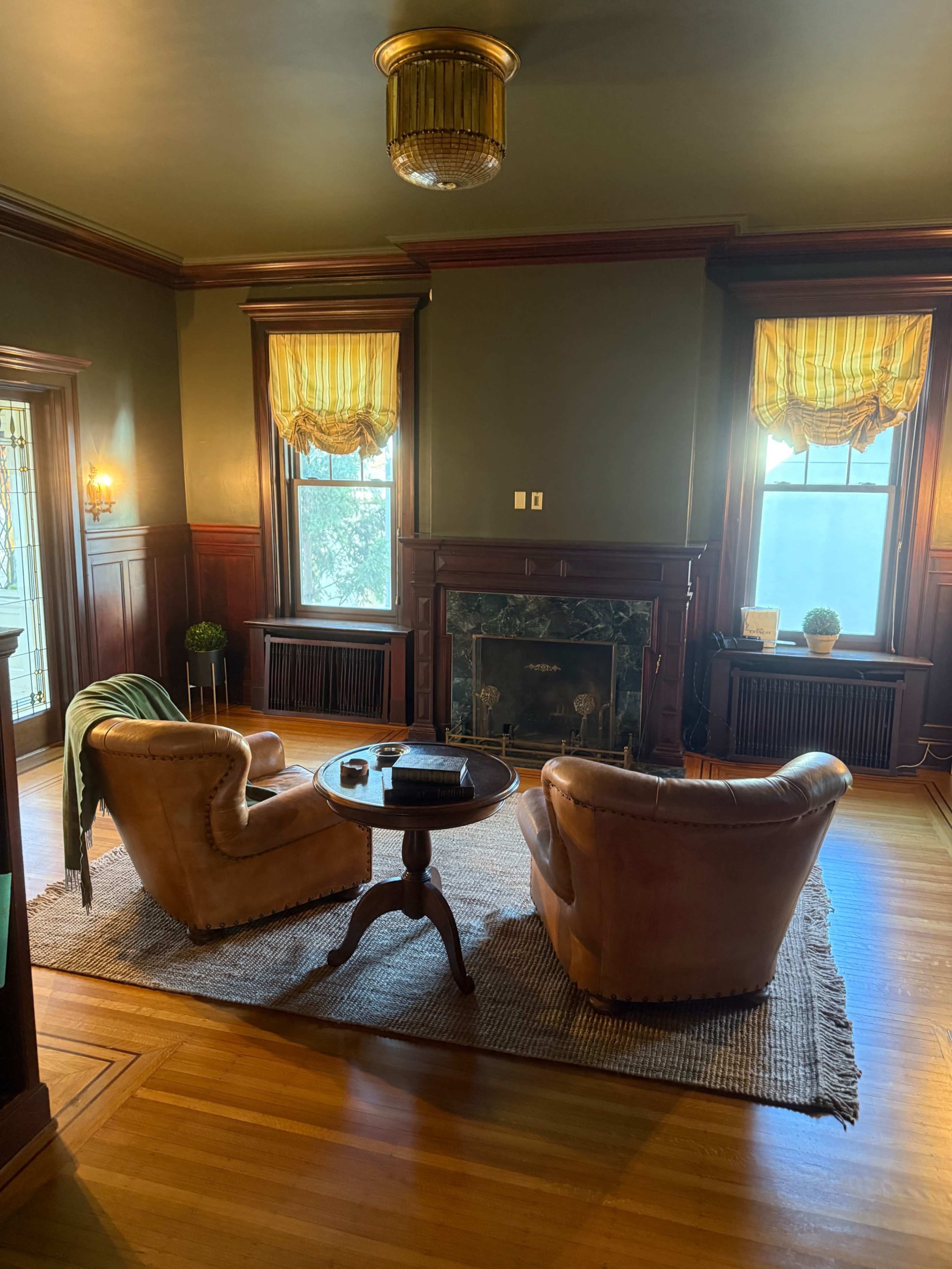 The image shows a cozy room with two armchairs facing a round table, a fireplace in the background, and large windows adorned with yellow curtains.
