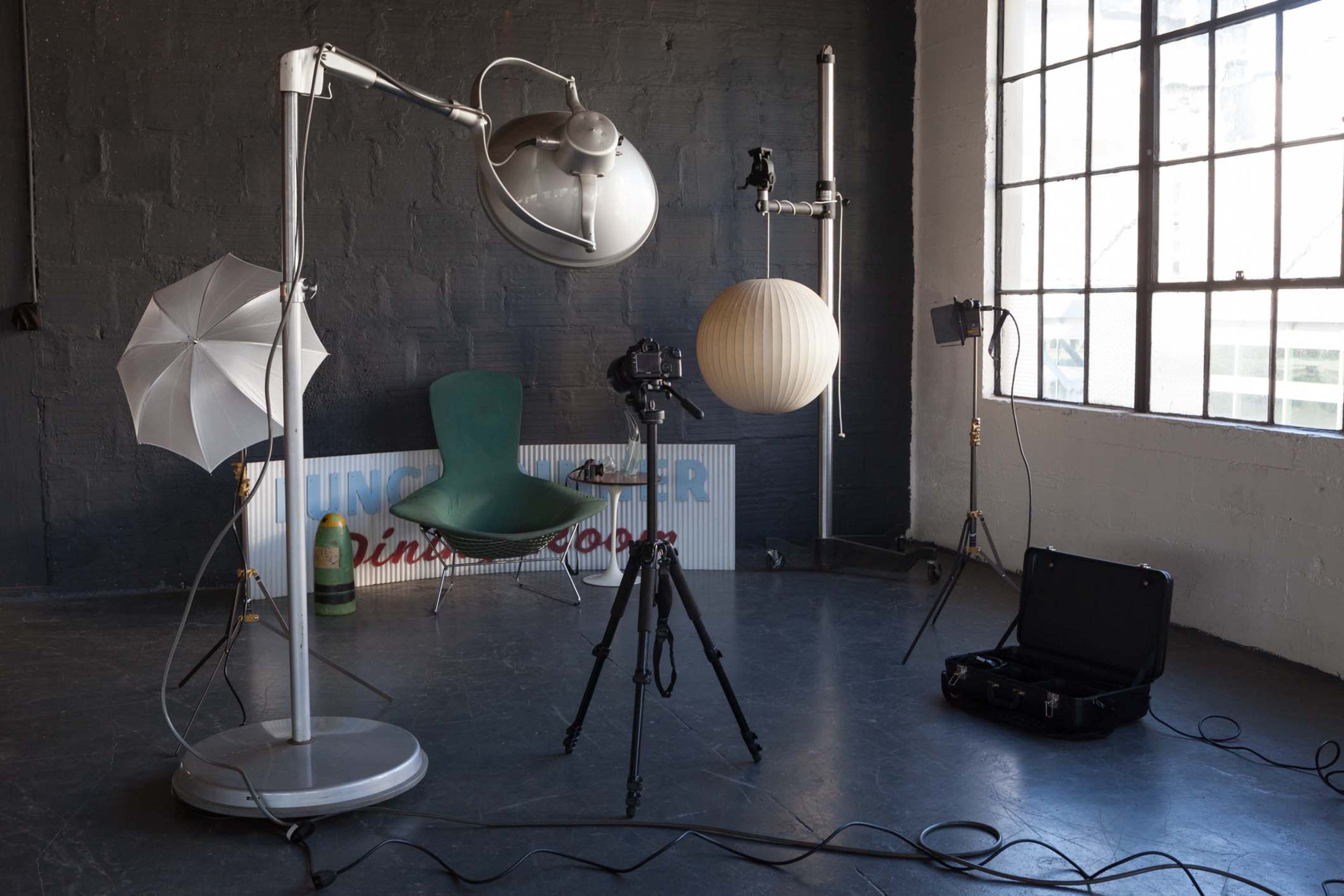 A photography studio setup with a camera on a tripod, a large light umbrella, and a round paper lamp hanging from the ceiling.