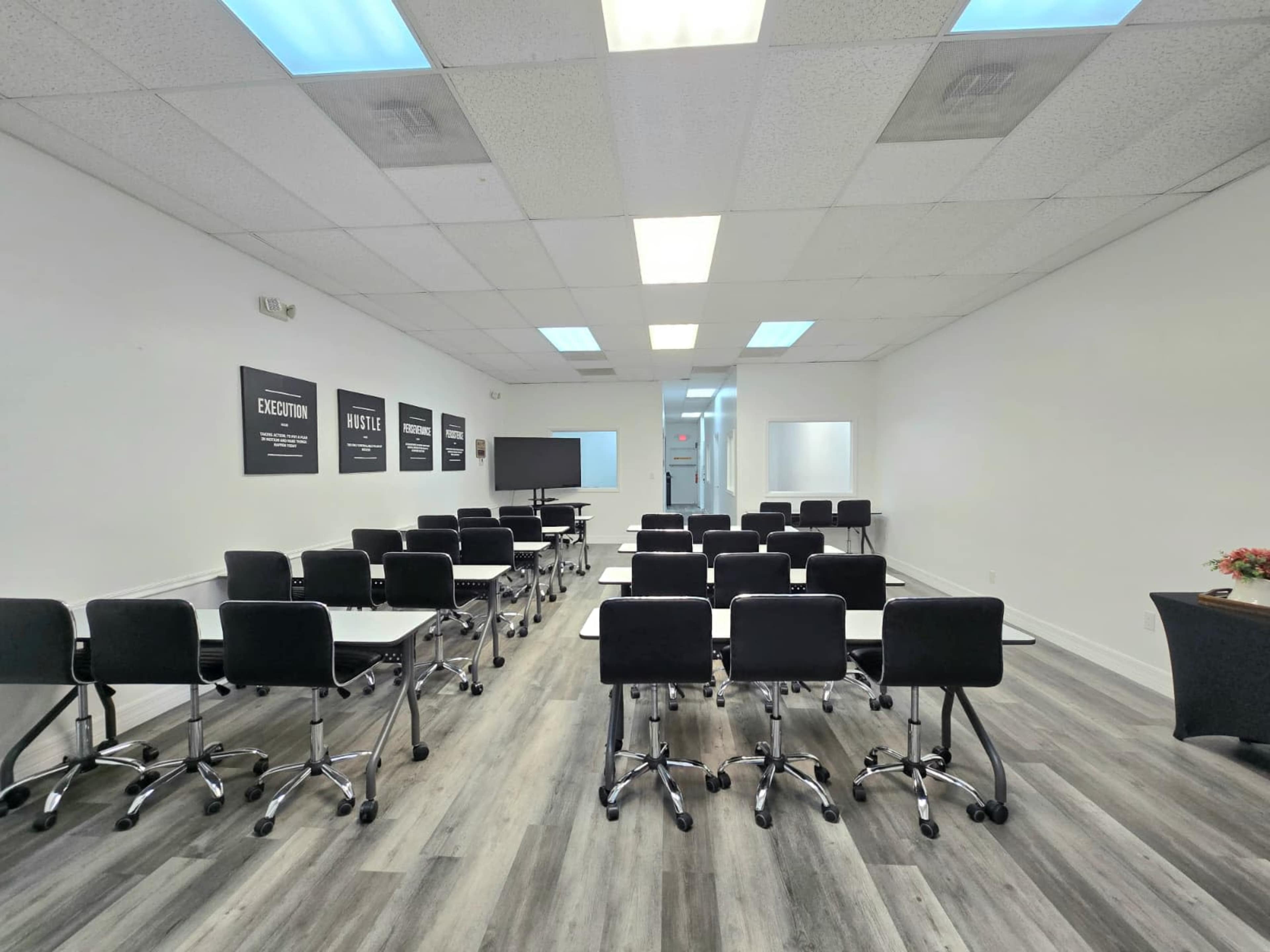 The image shows a conference room with rows of black chairs and tables arranged for a meeting, along with a screen at the front and wall posters.