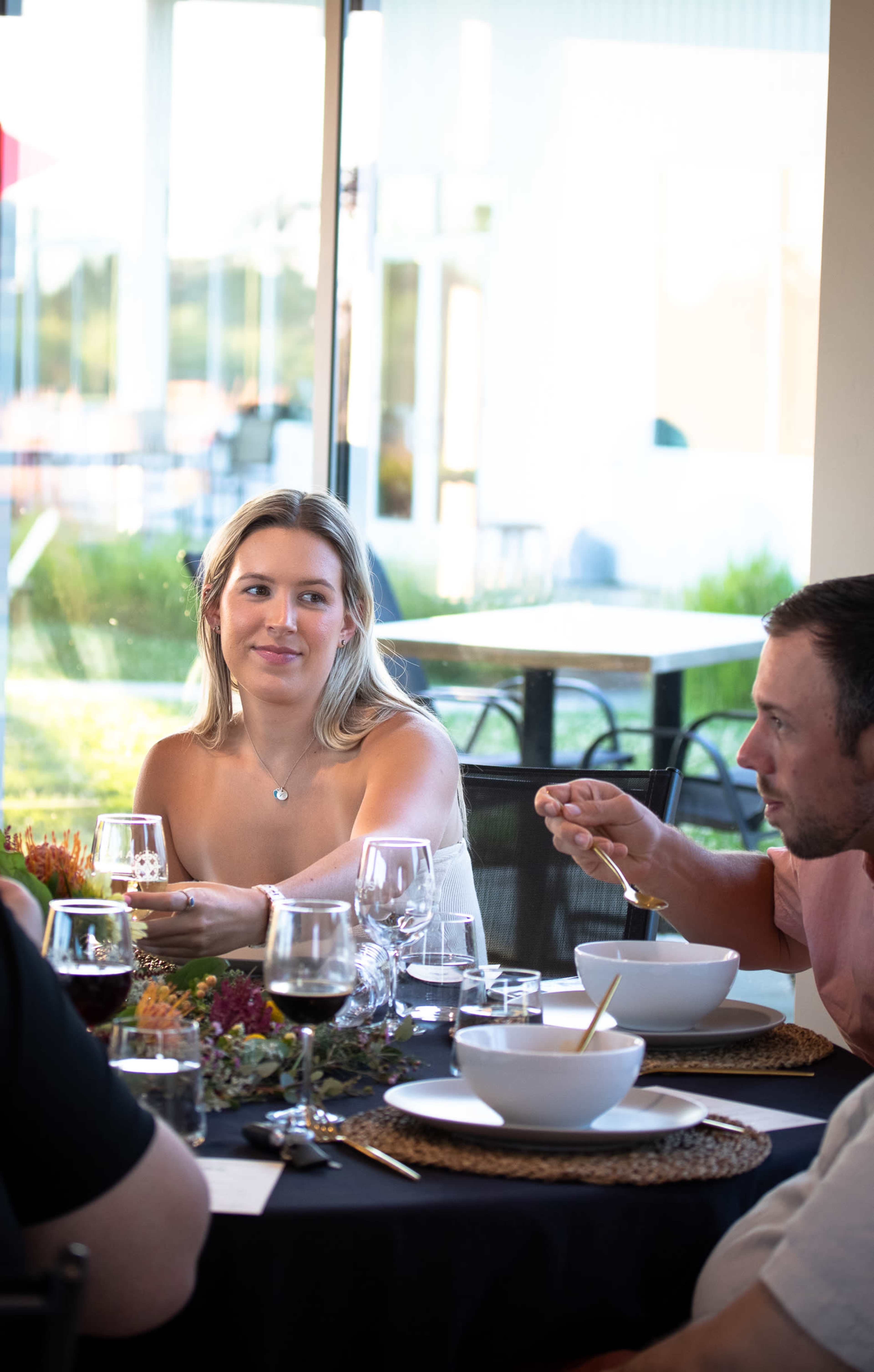 A woman and a man are seated at a dining table with glassware and bowls, engaged in conversation in a bright restaurant setting.