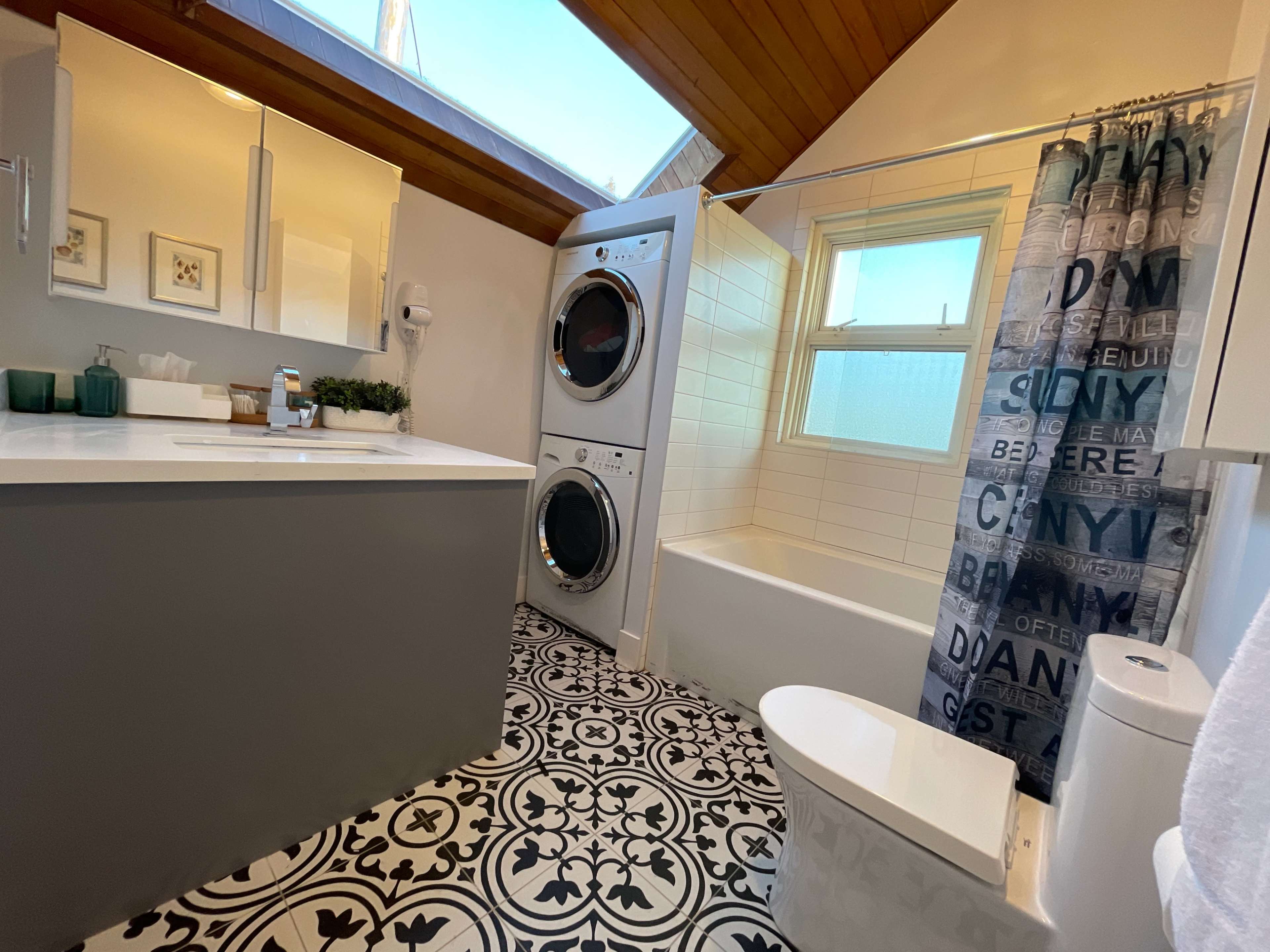 The bathroom features a bathtub with a shower, stacked laundry machines, a modern vanity with a sink, and patterned tile flooring.