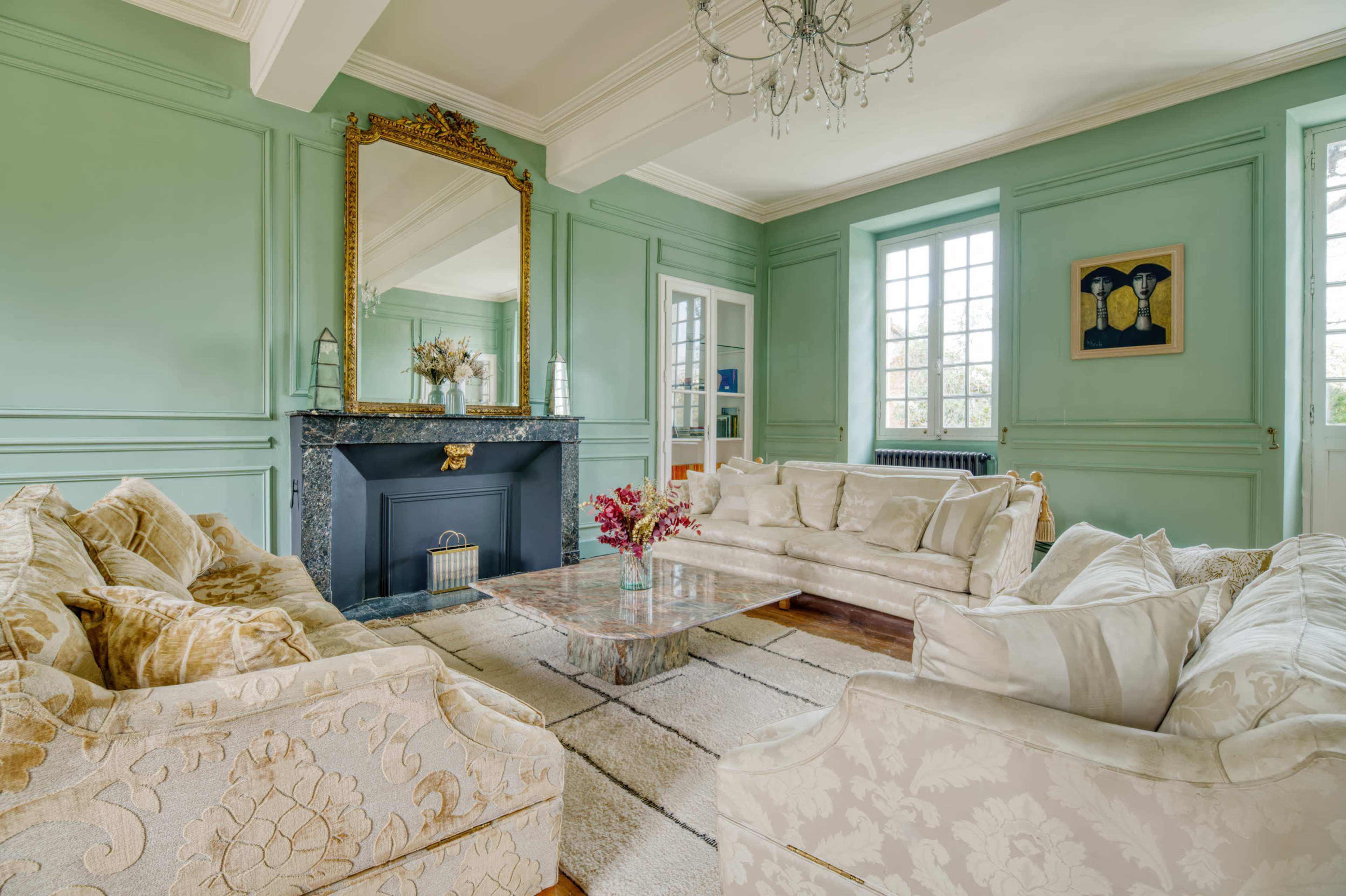 The image shows a spacious living room with light green walls, a marble fireplace, and two upholstered sofas arranged around a glass coffee table.