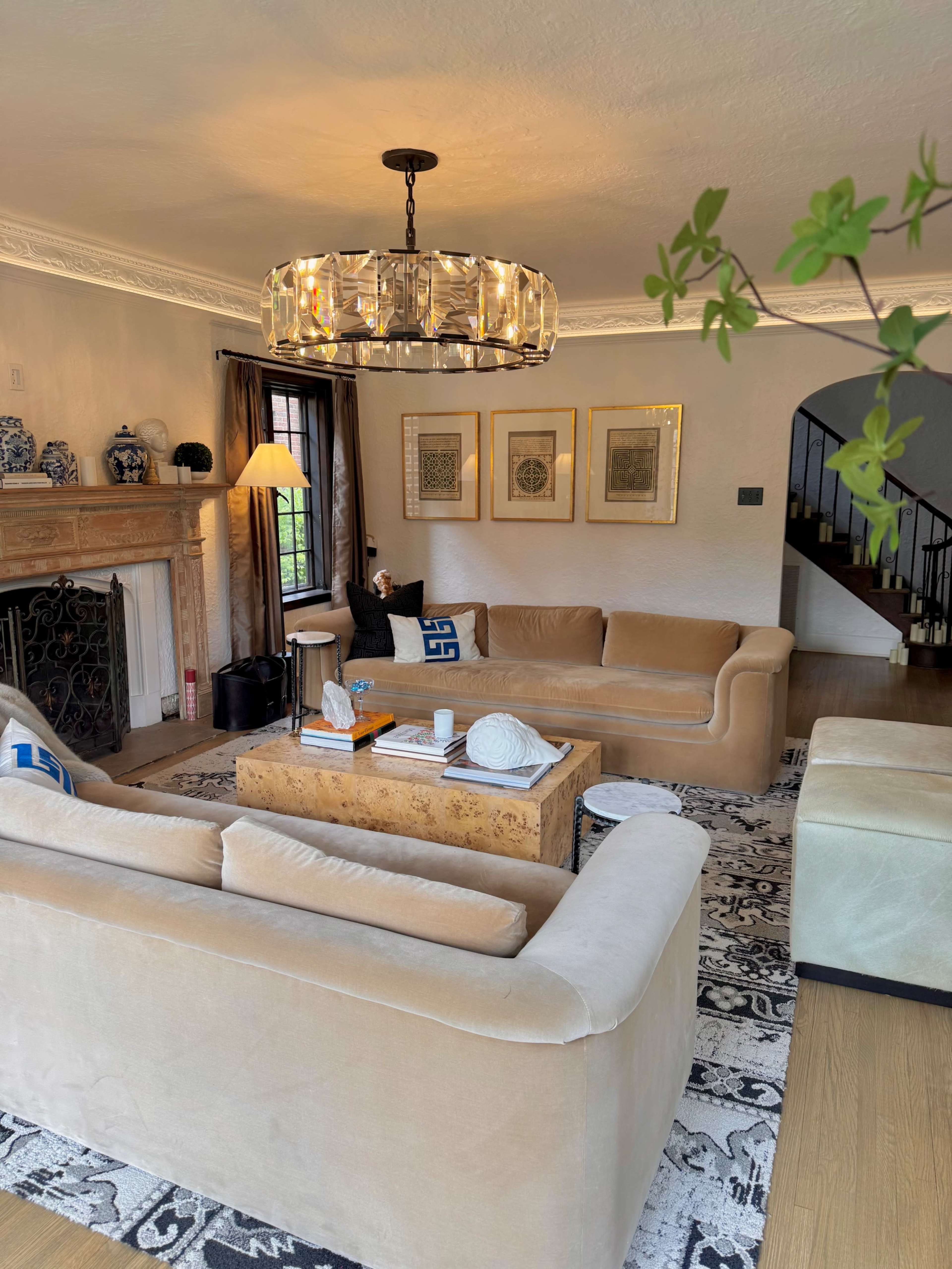 The image shows a cozy living room featuring two beige sofas, a wooden coffee table, framed artworks on the wall, and a chandelier overhead.