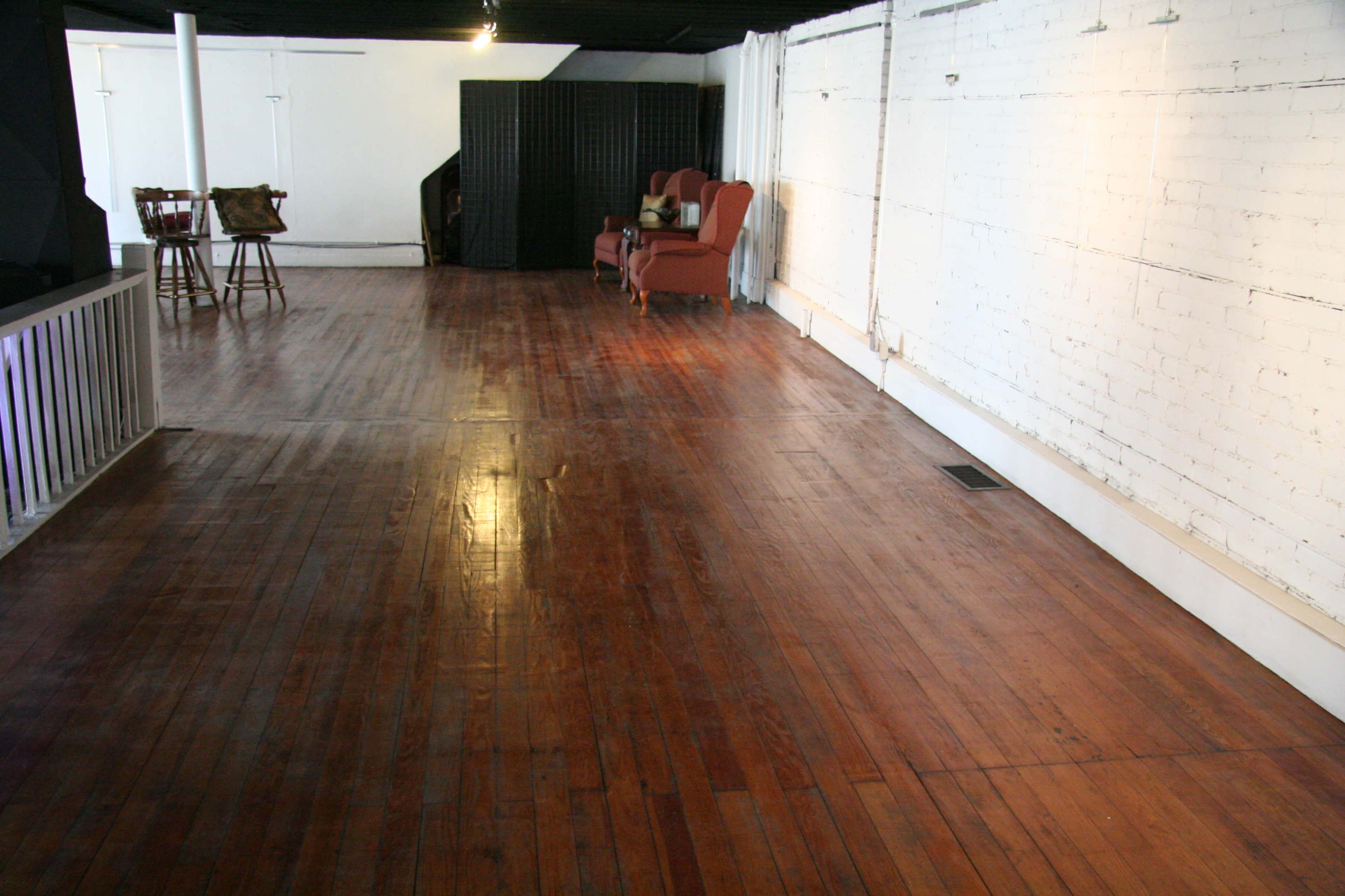 A spacious room with polished wooden floors, a white brick wall, and a few pieces of furniture including a brown couch and a bar setup at one end.