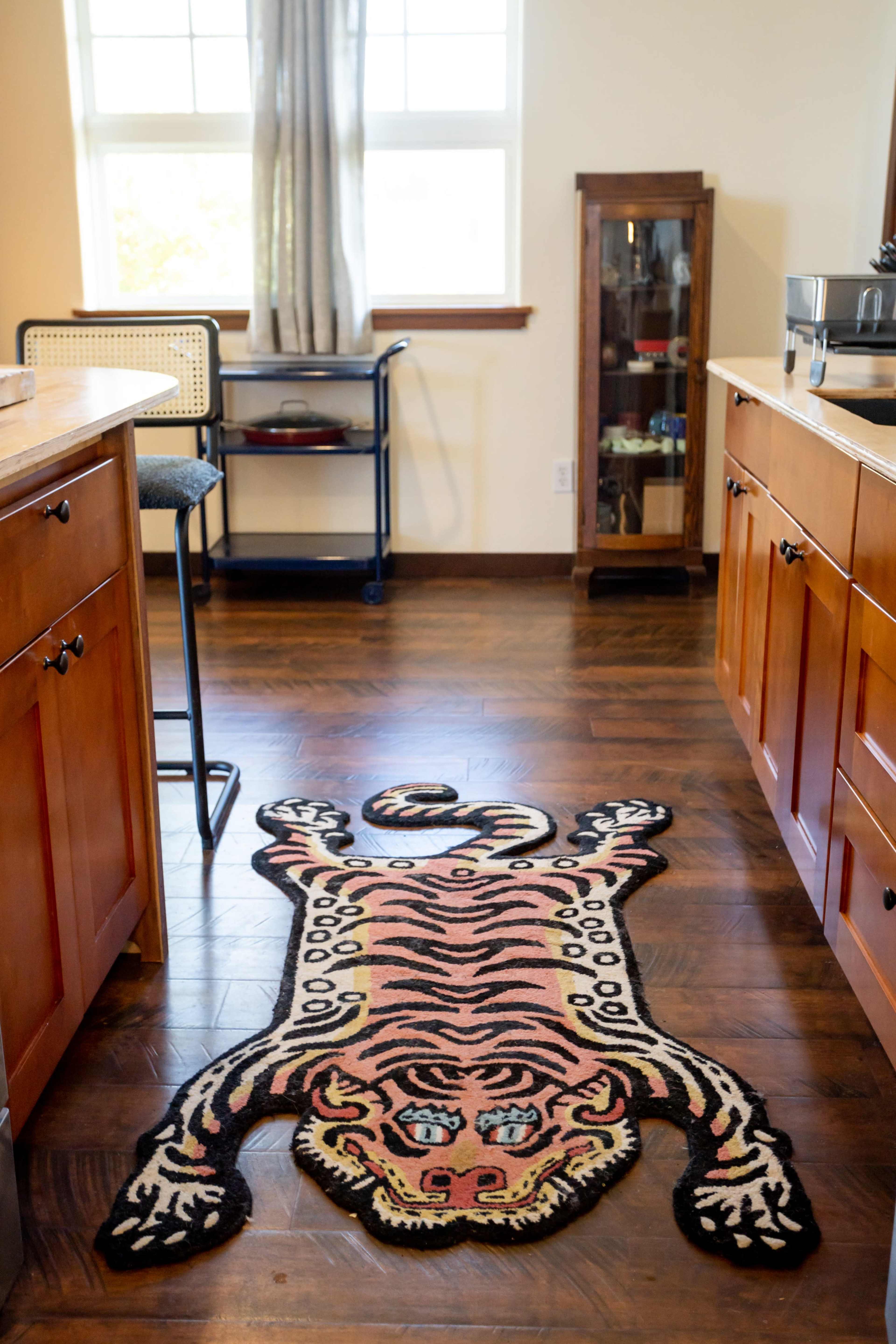 A tiger-patterned rug lays on the wooden floor of a kitchen, positioned between wooden cabinets and a bar stool.