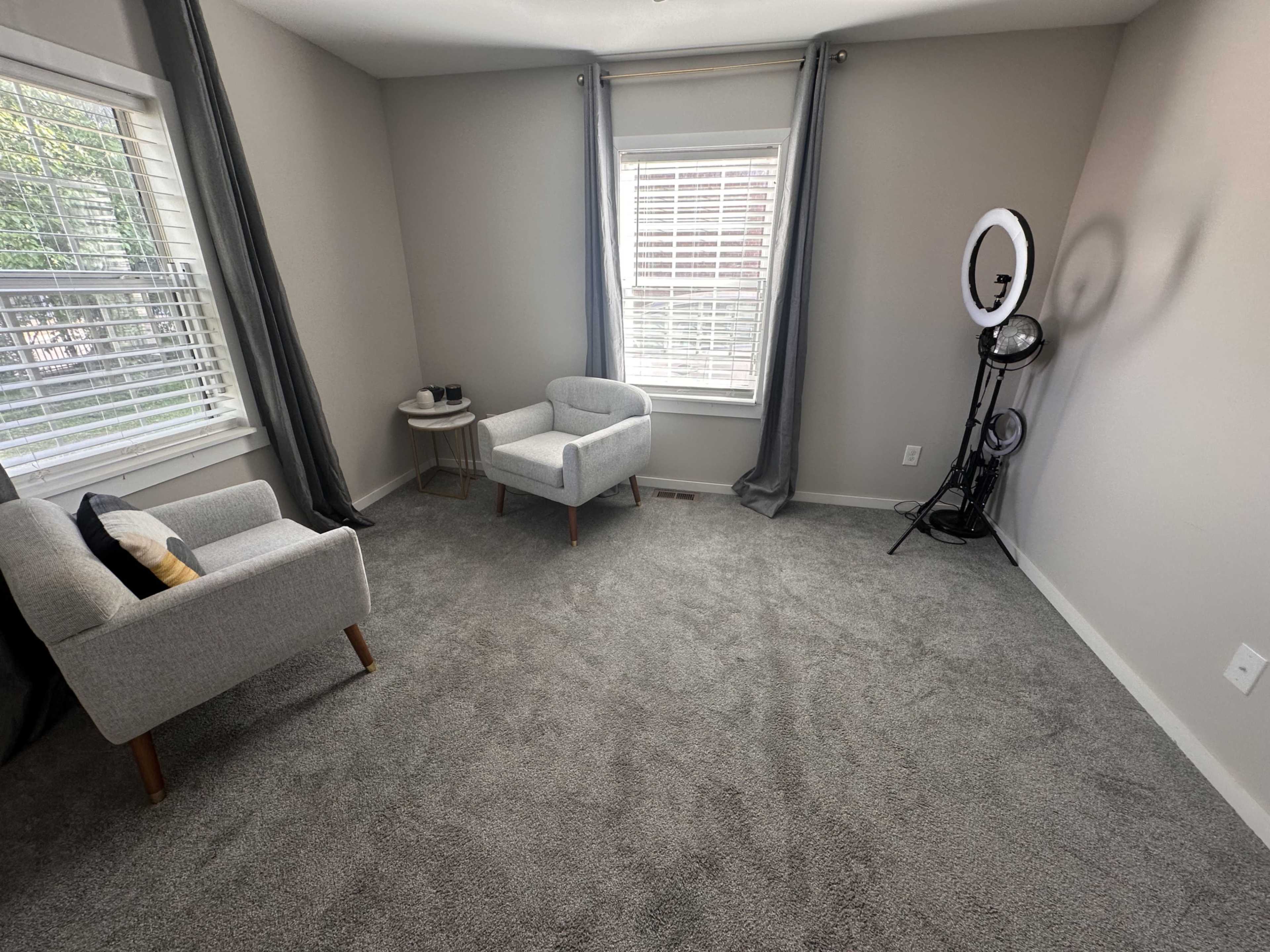 The image shows a minimalist room with gray carpet, two light gray chairs, a small round table, and a ring light set against a wall with large windows.