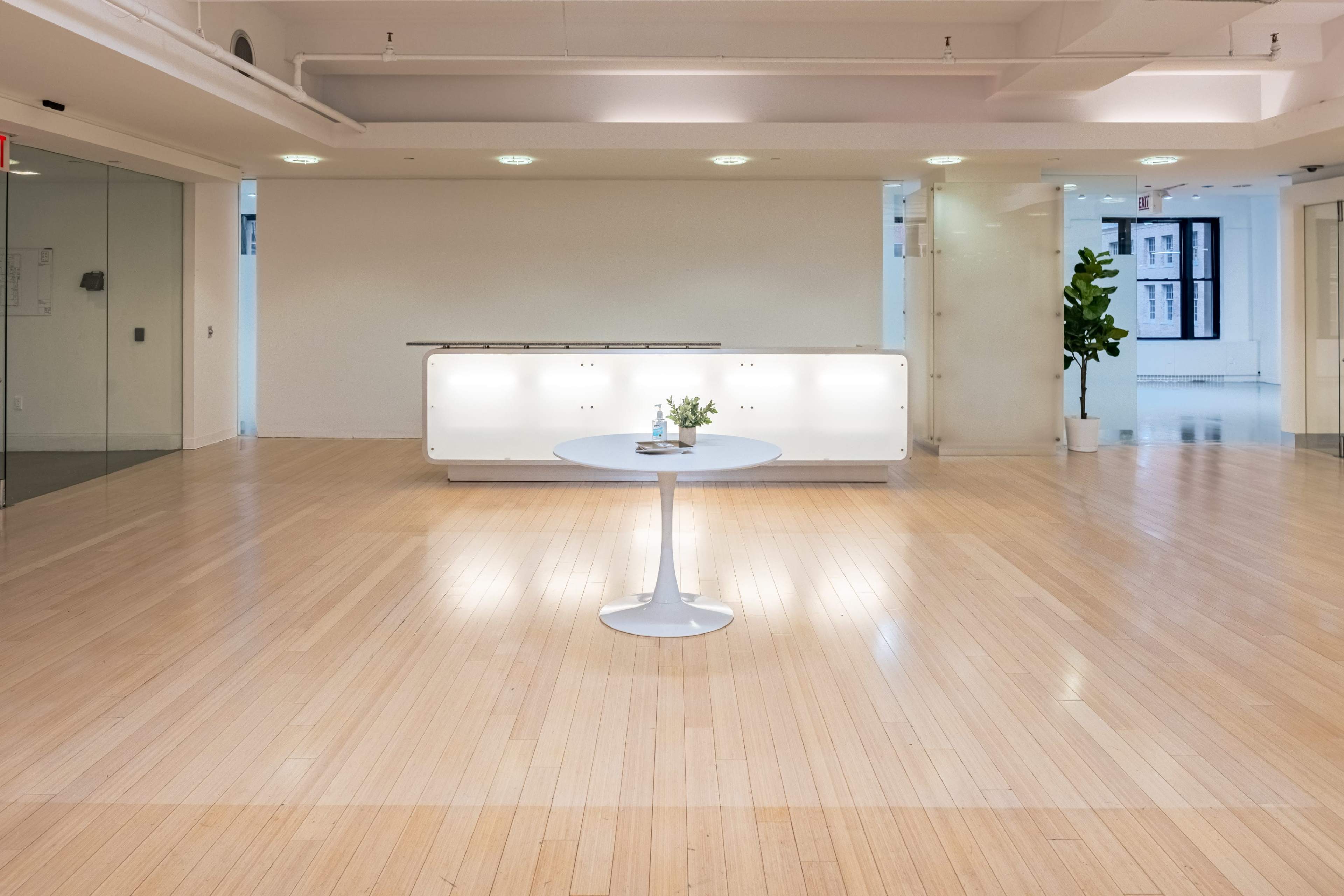 The image shows a spacious, bright room with light wooden flooring, a small round table in the center, and a modern reception desk at the back.