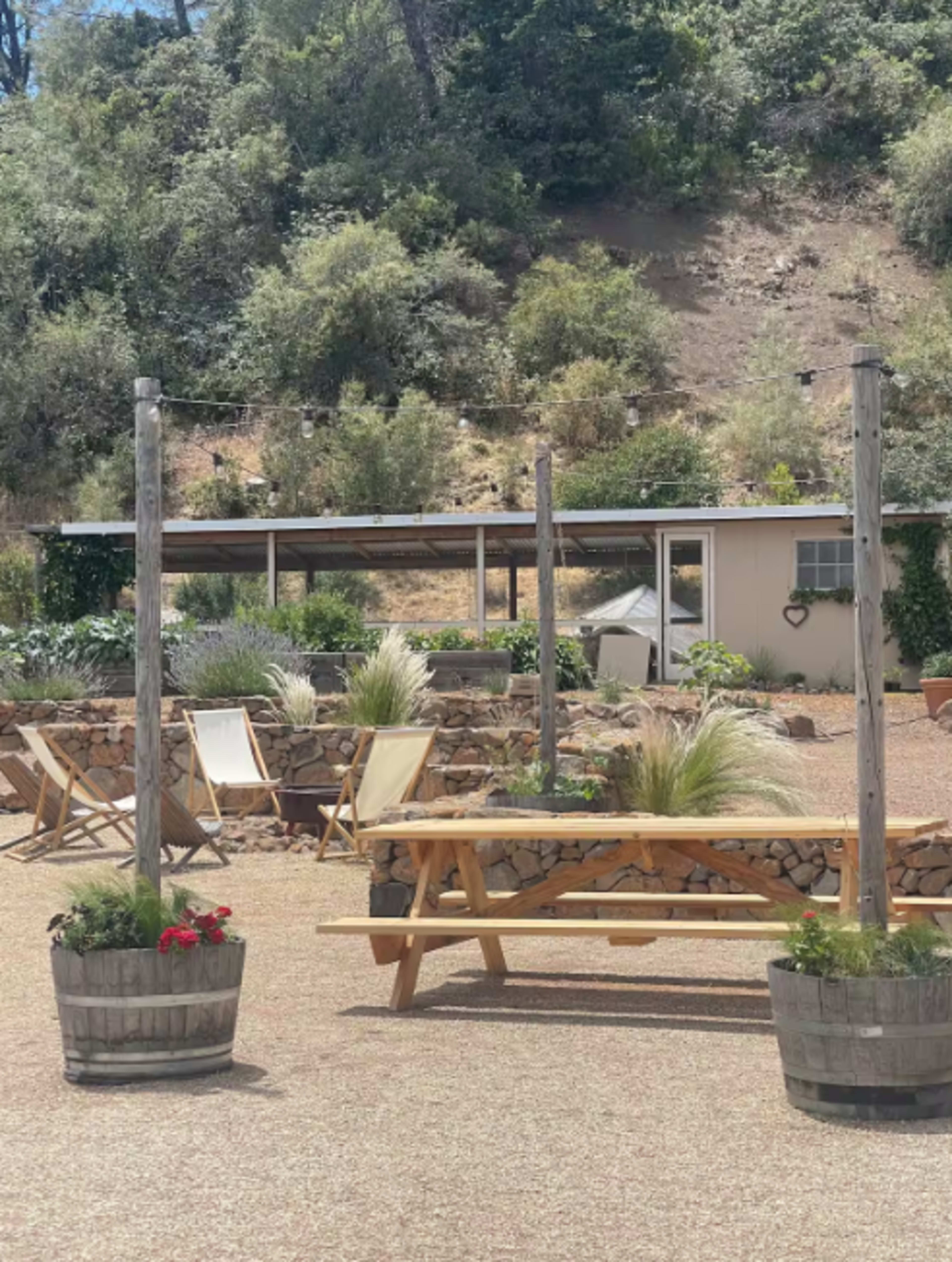 The image shows a rustic outdoor area featuring a wooden picnic table, chairs, and planters, set against a hillside covered with greenery.