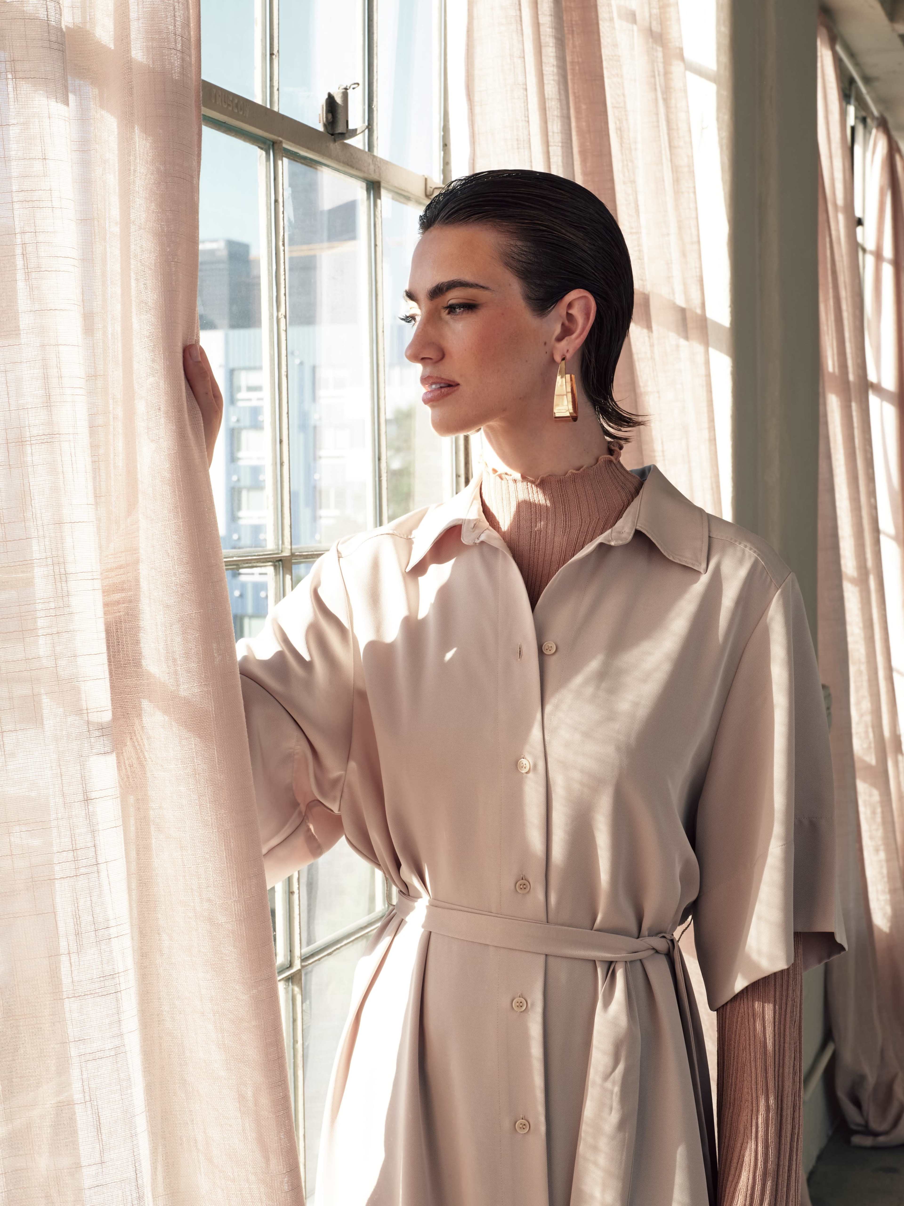 A woman in a light, beige dress stands by a large window, opening sheer curtains to let in natural light.