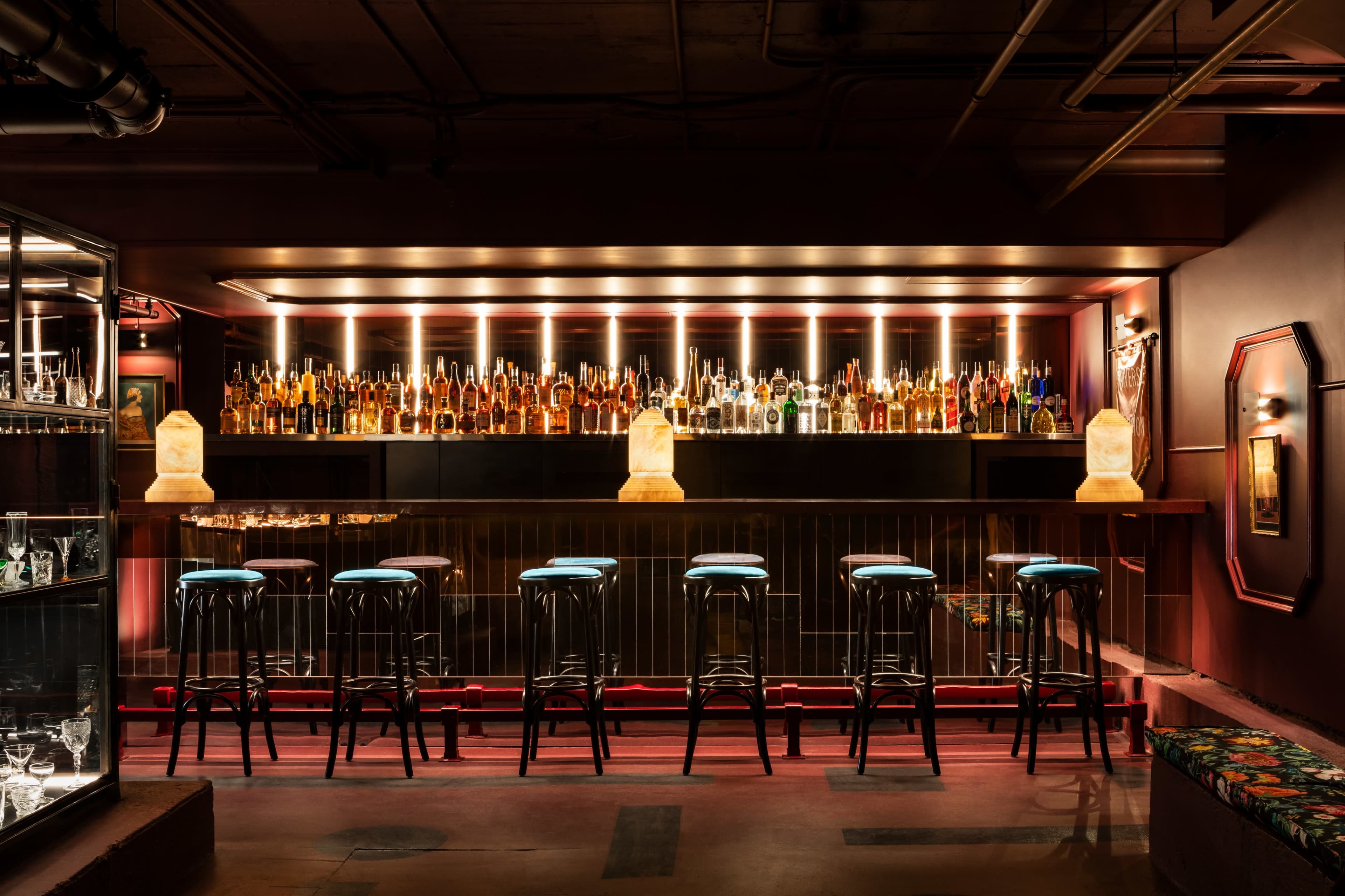 A dimly lit bar features a long counter with illuminated shelves filled with bottles and several high stools lined up in front.