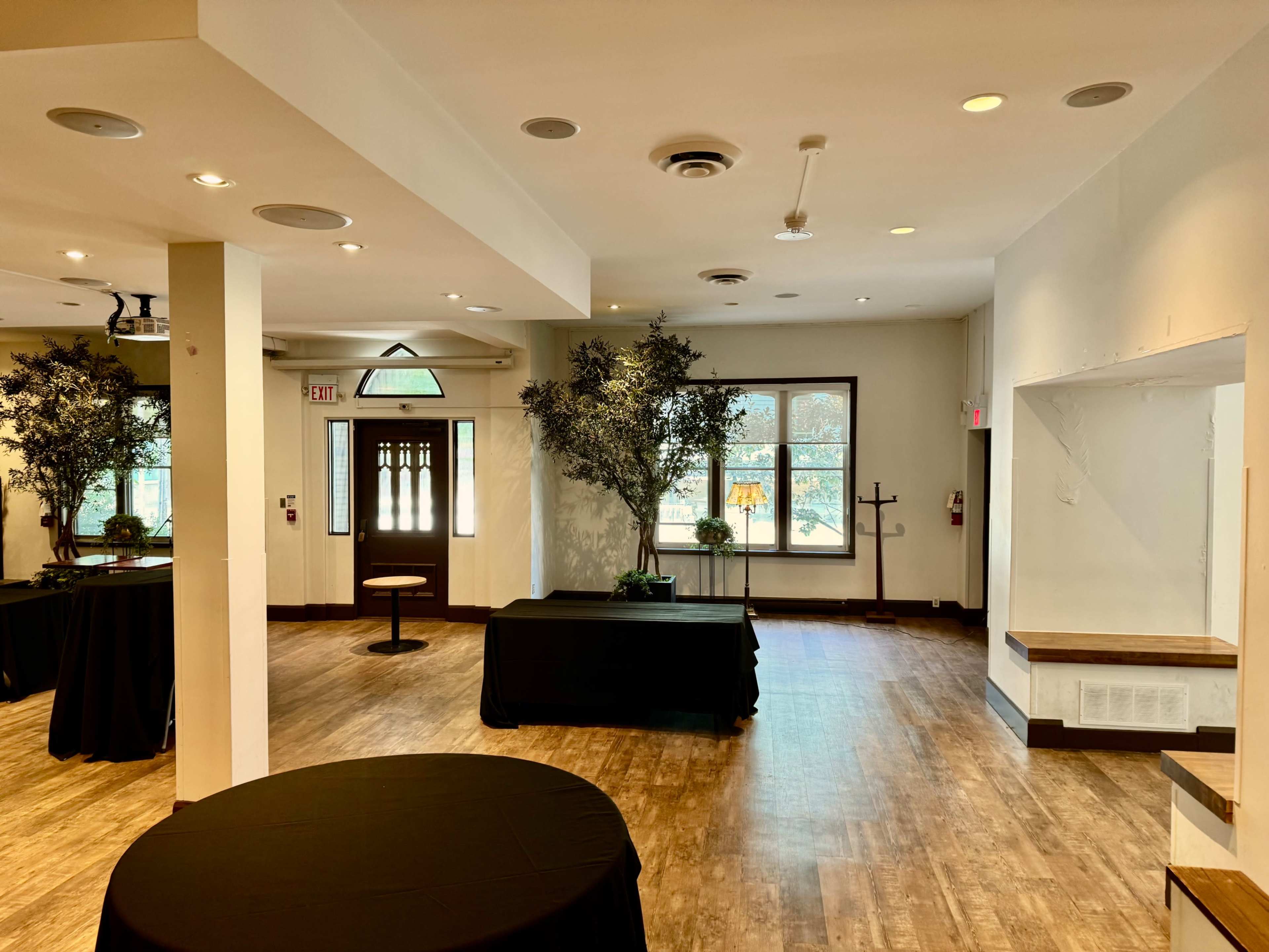 The image shows an indoor event space with wooden flooring, a few tables covered in black cloth, and large windows allowing natural light to enter.