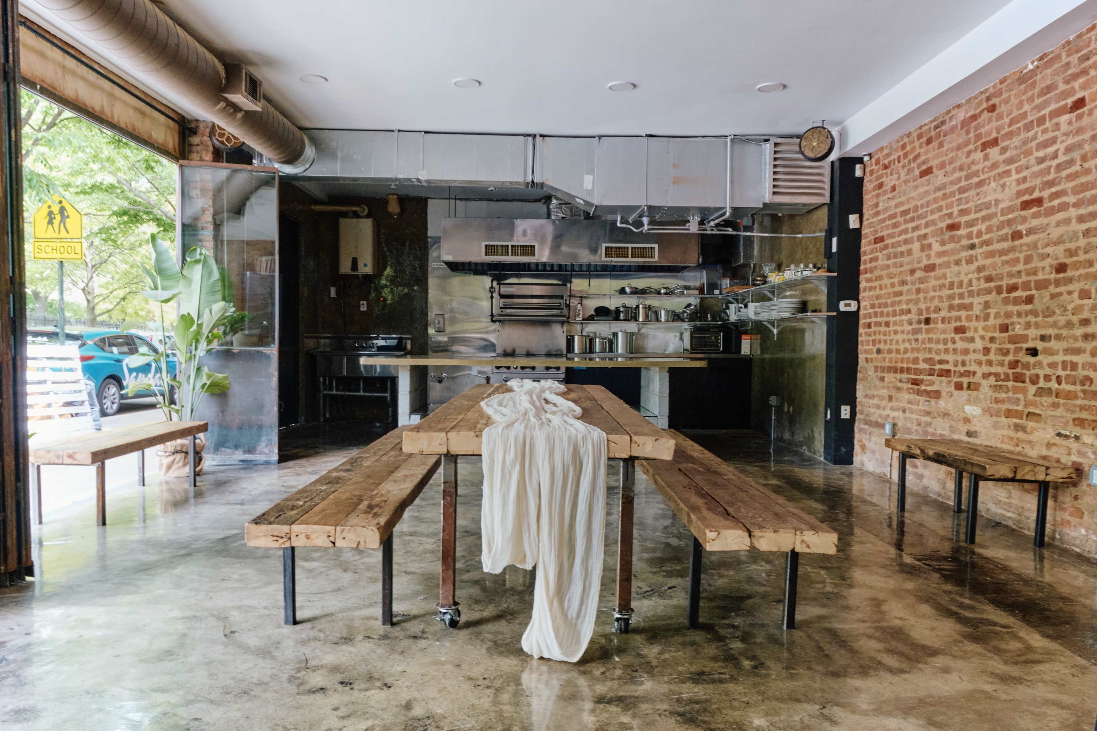 An empty restaurant interior with a long wooden table at the center, surrounded by benches, and a visible kitchen area in the background.