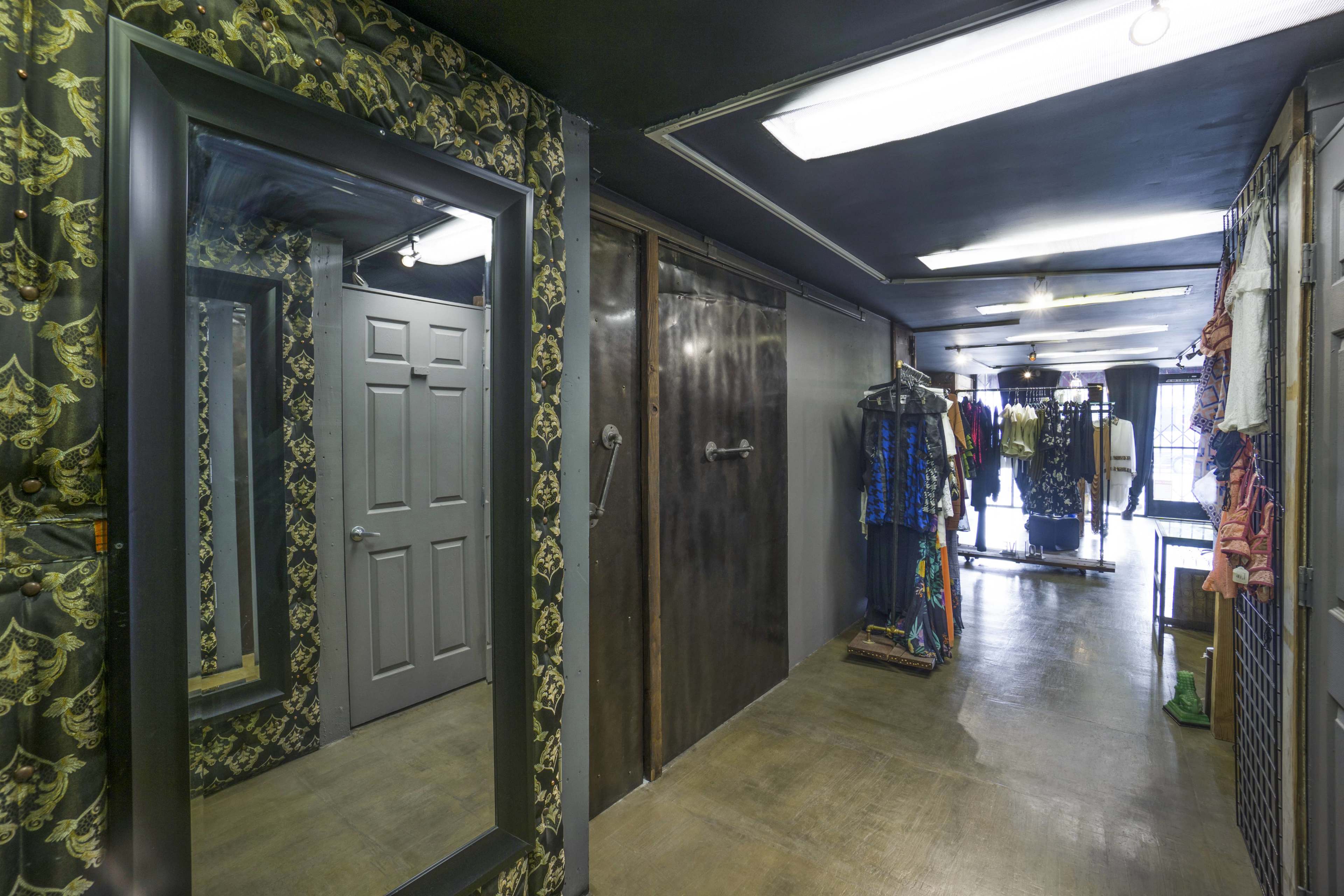 A narrow boutique interior with a large mirror, a gray door, a metallic sliding door, and clothing racks along the walls.