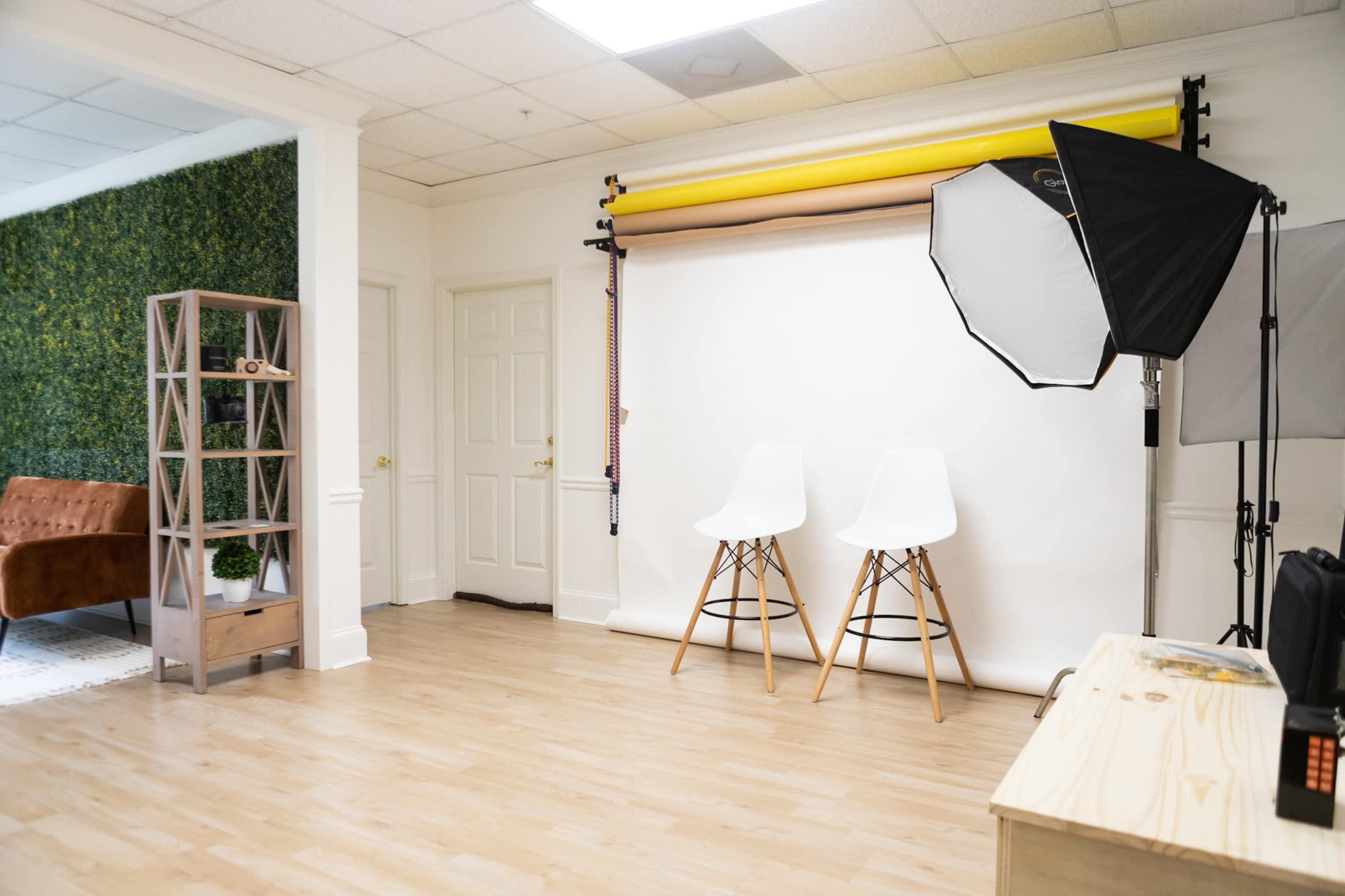 The image shows a bright studio space with two white chairs in front of a beige backdrop and a softbox light set up for photography.