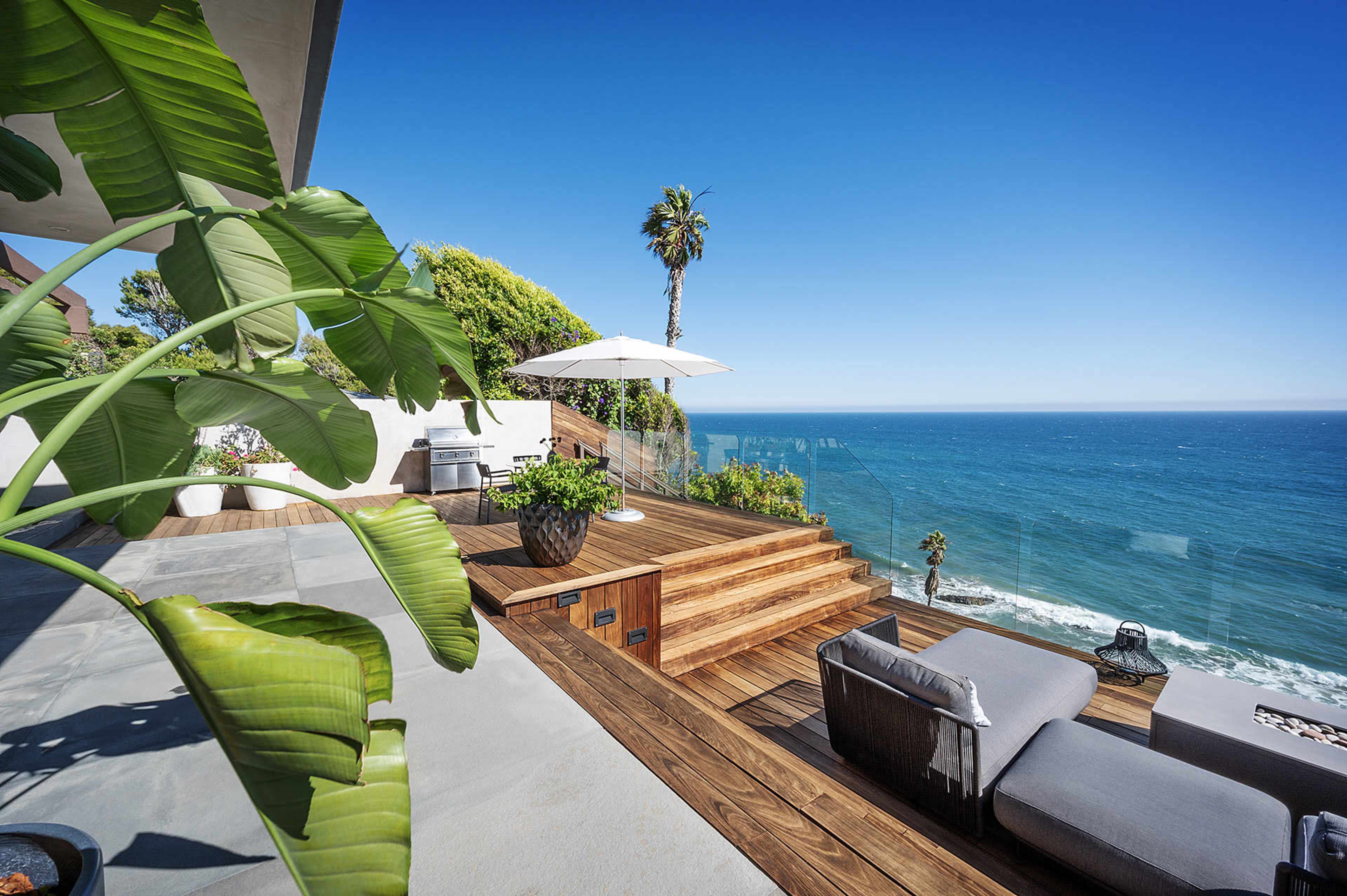 A wooden deck with outdoor seating and a large umbrella overlooks the ocean under a clear blue sky.