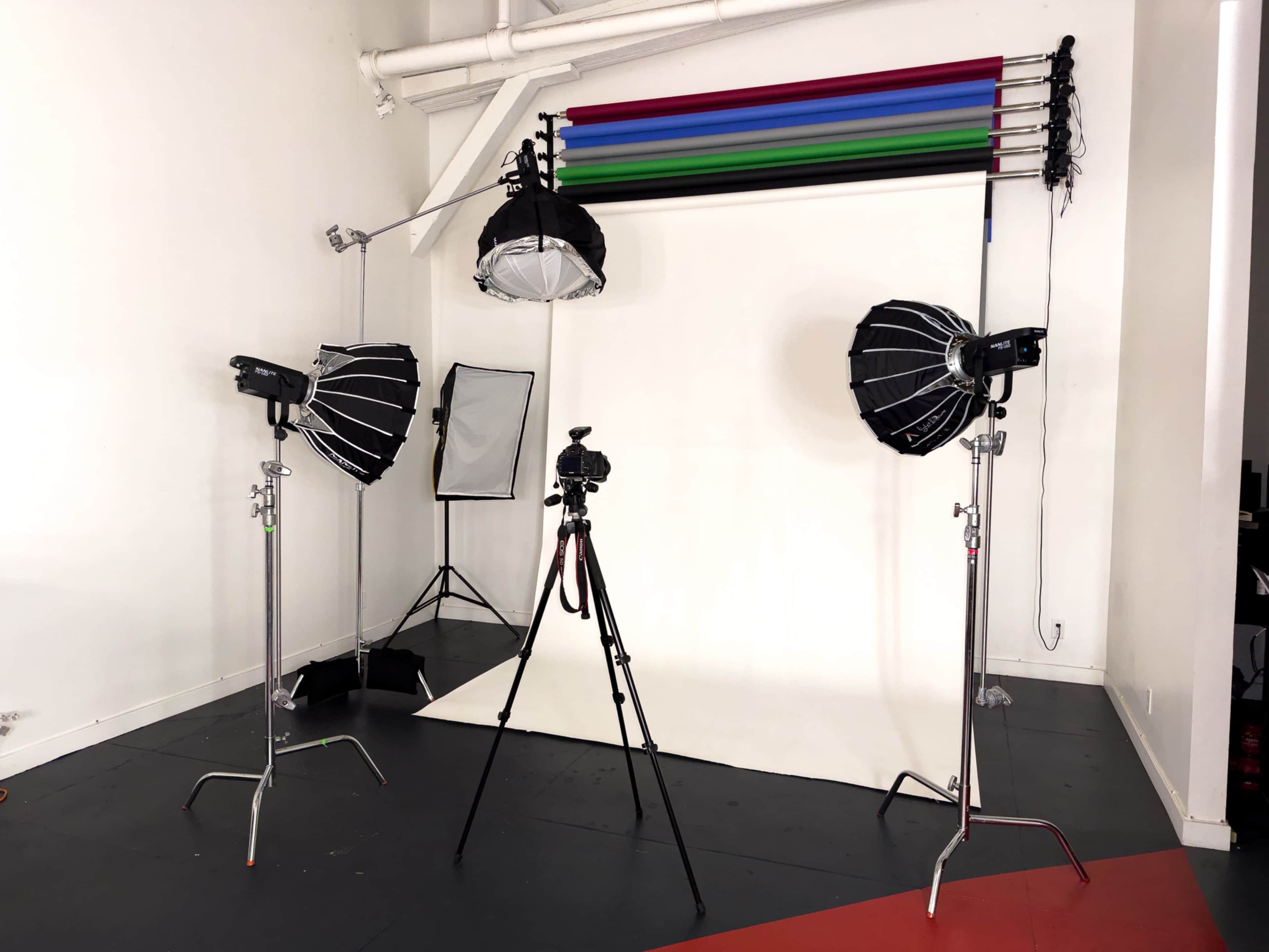 The image shows a photography studio set up with two softbox lights, a camera on a tripod, and a backdrop with colored strips.