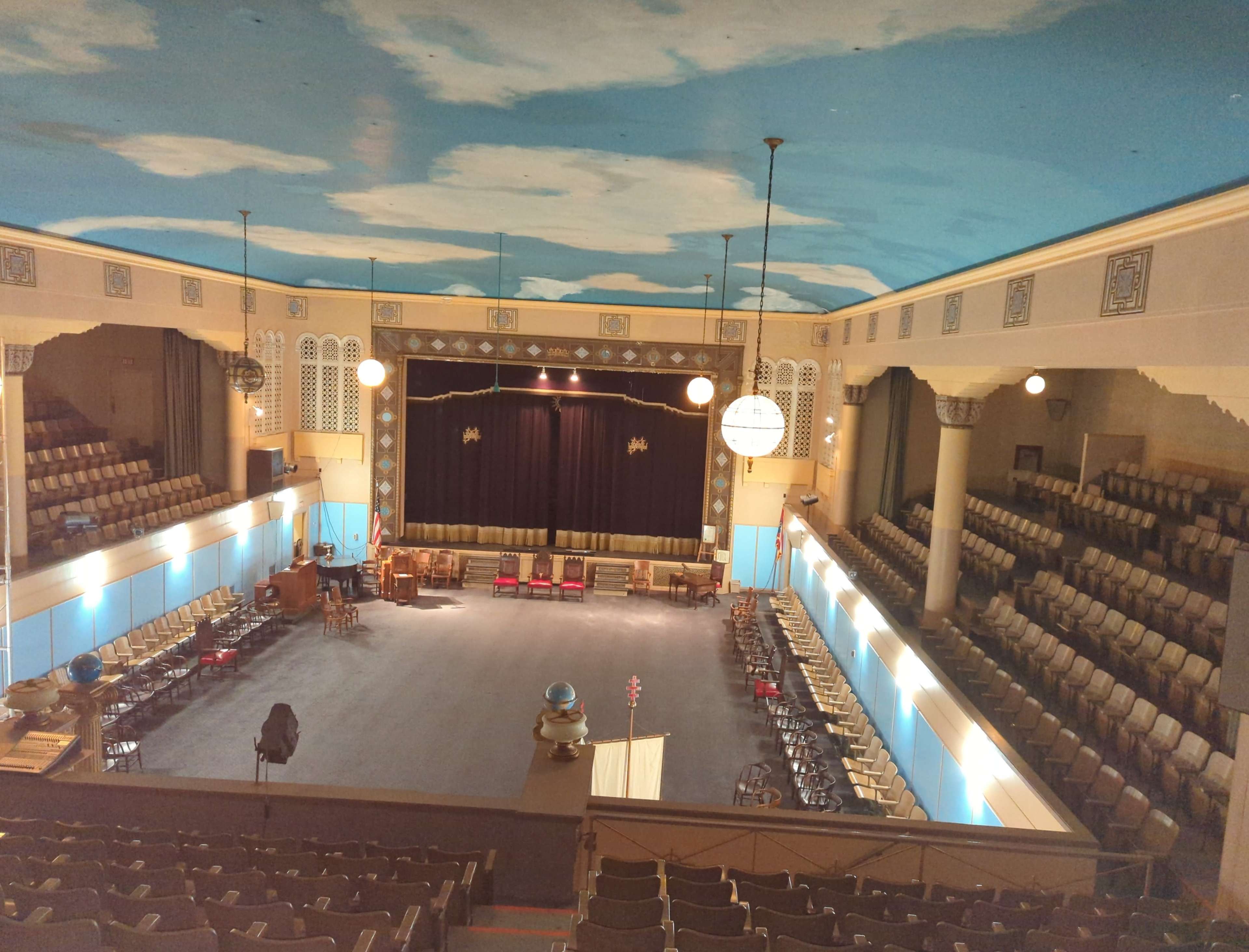 An empty theater with tiered seating, stage curtains drawn, and a painted sky ceiling.