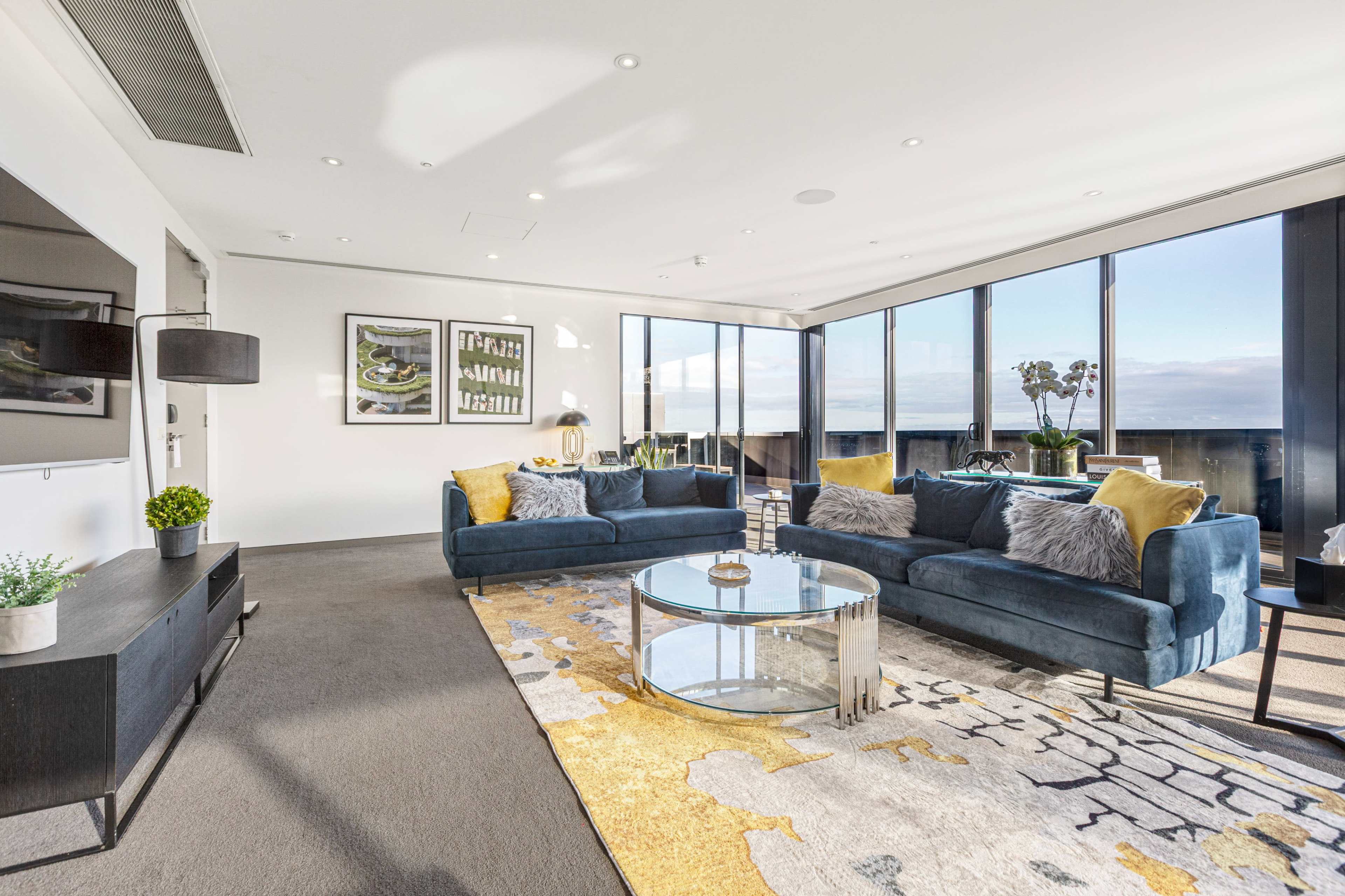 The image shows a modern living room featuring two blue sofas, a circular glass coffee table, and large windows providing a view of the outdoors.