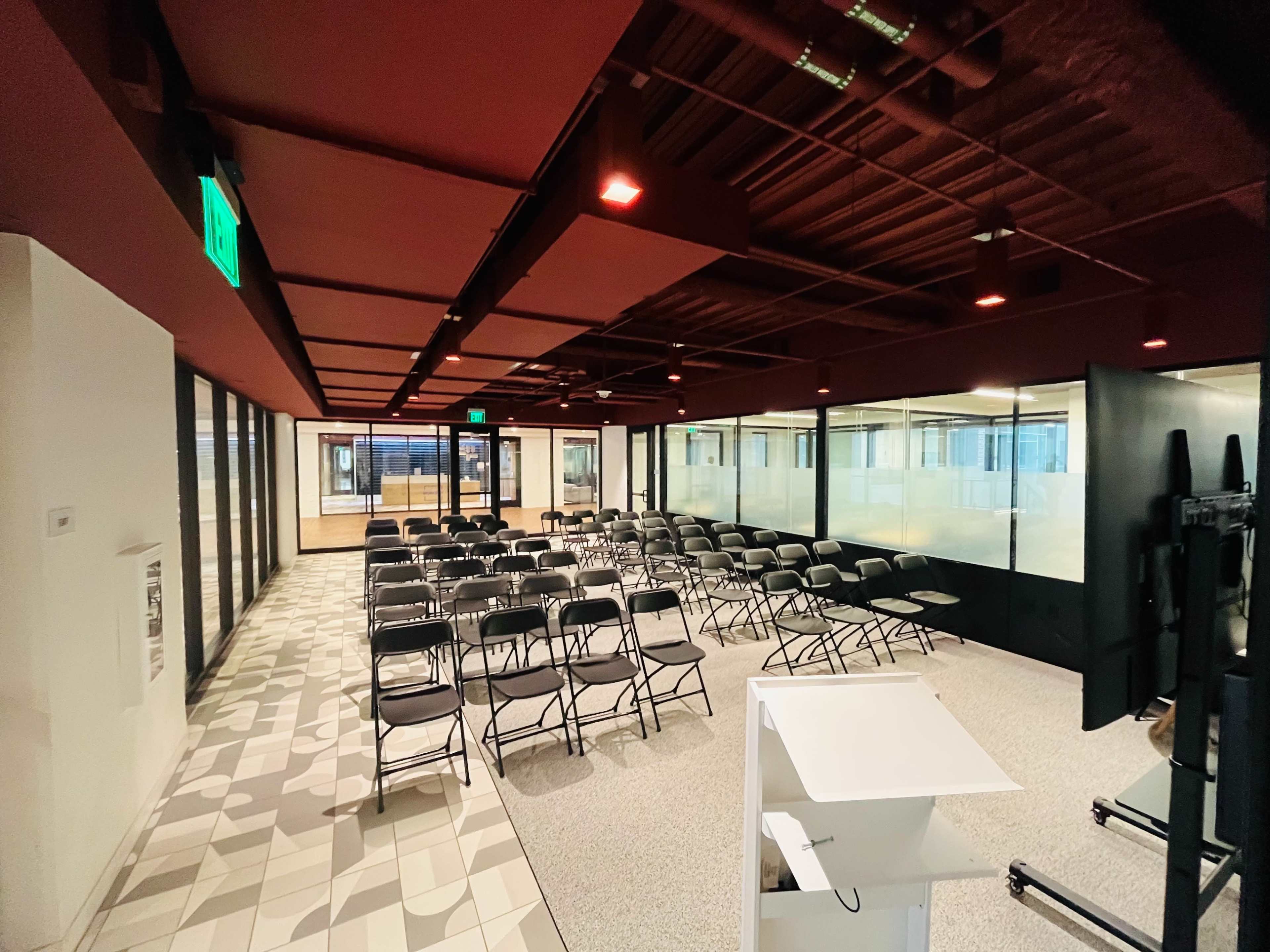 A spacious meeting room with rows of black chairs arranged on a patterned floor, surrounded by glass walls and equipped with a presentation podium.