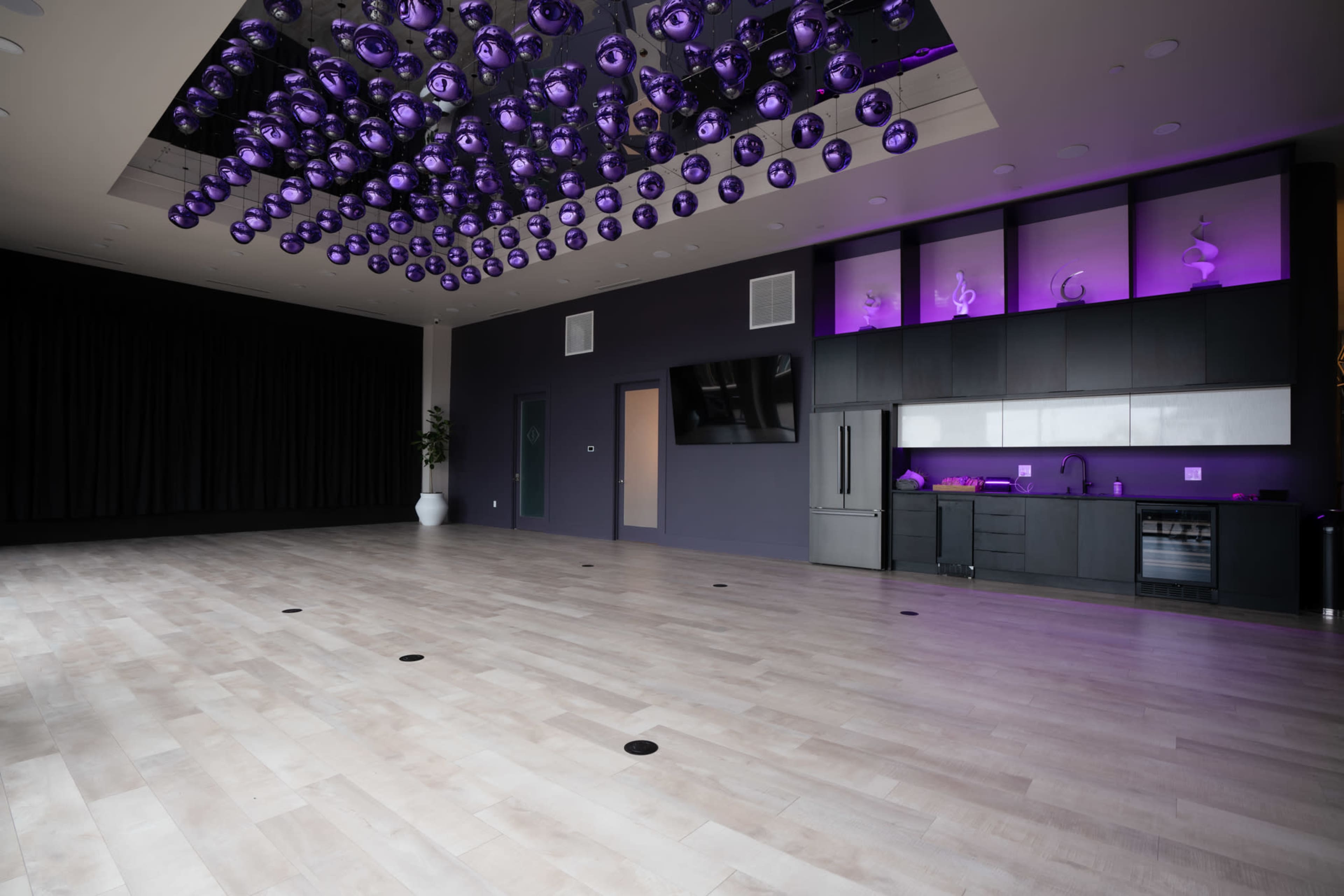 The image depicts a spacious indoor room with light wood flooring, a dark feature wall, and a ceiling adorned with an array of purple light fixtures.