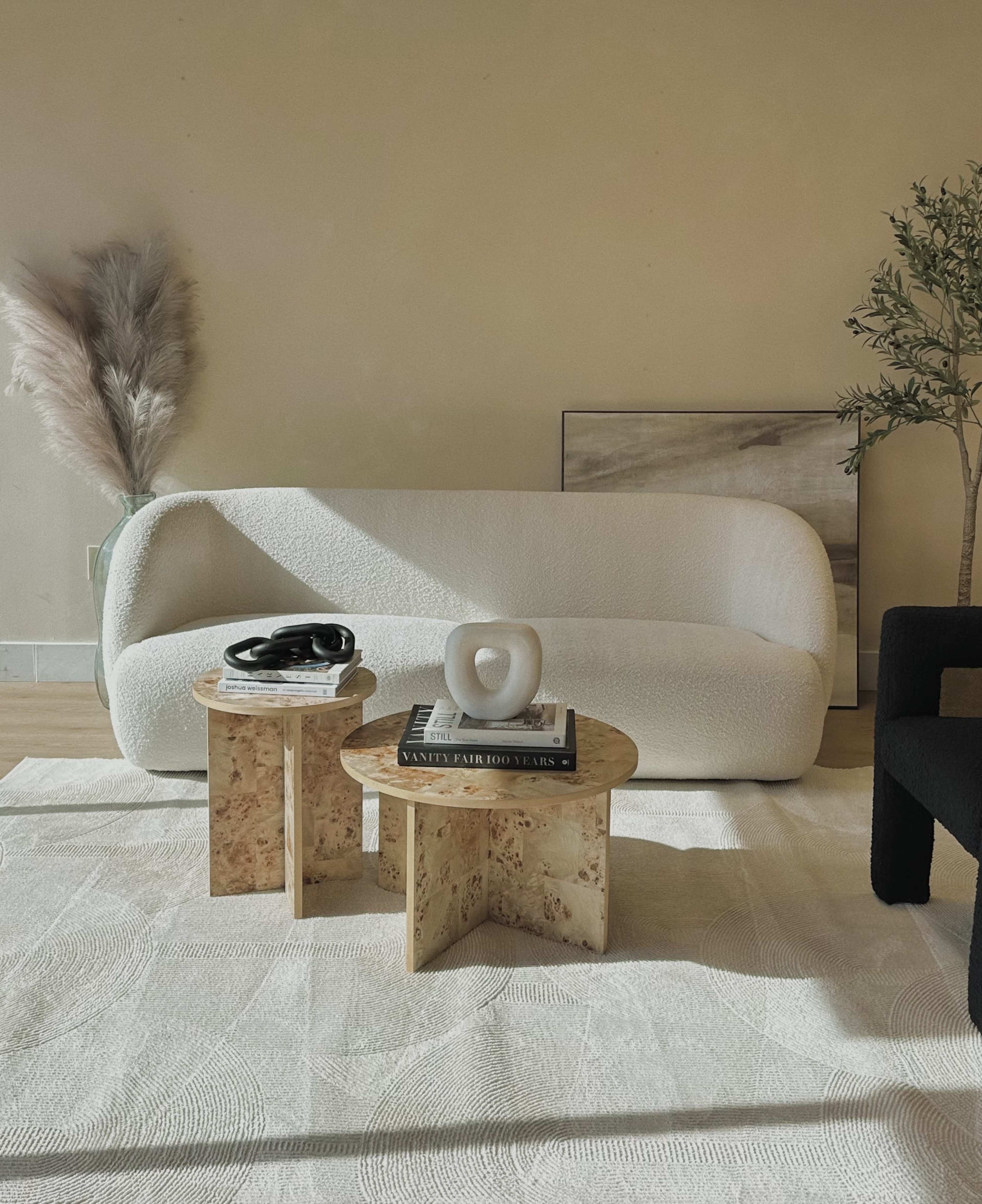 The image shows a minimalist living room setting featuring a white curved sofa, two round tables with light stone finishes, a small decorative sculpture, and a couple of decorative elements like a plant and artwork on the wall.