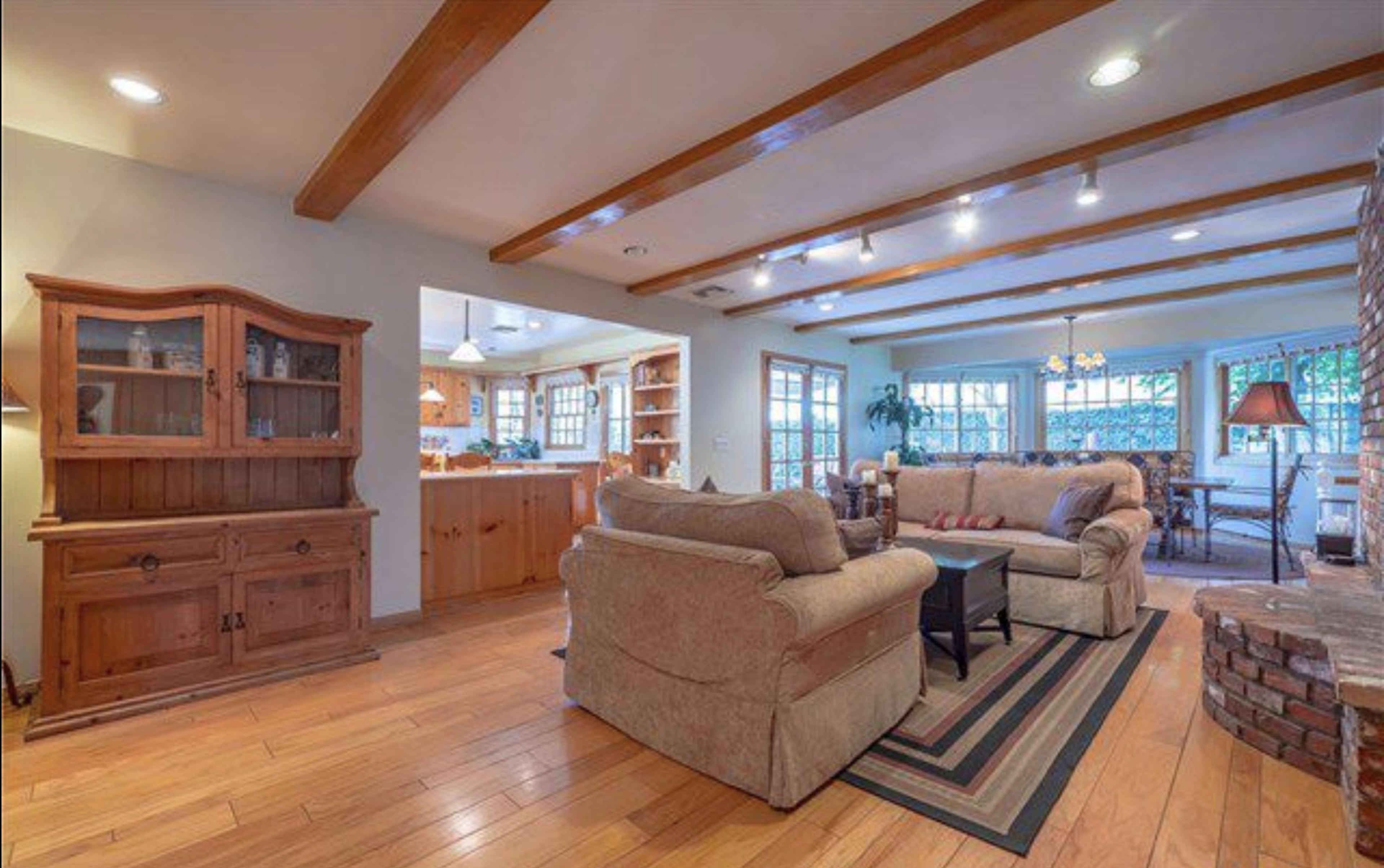 A living room features two sofas facing a black coffee table, with wooden beams on the ceiling and a view into a kitchen area with light wooden cabinetry.