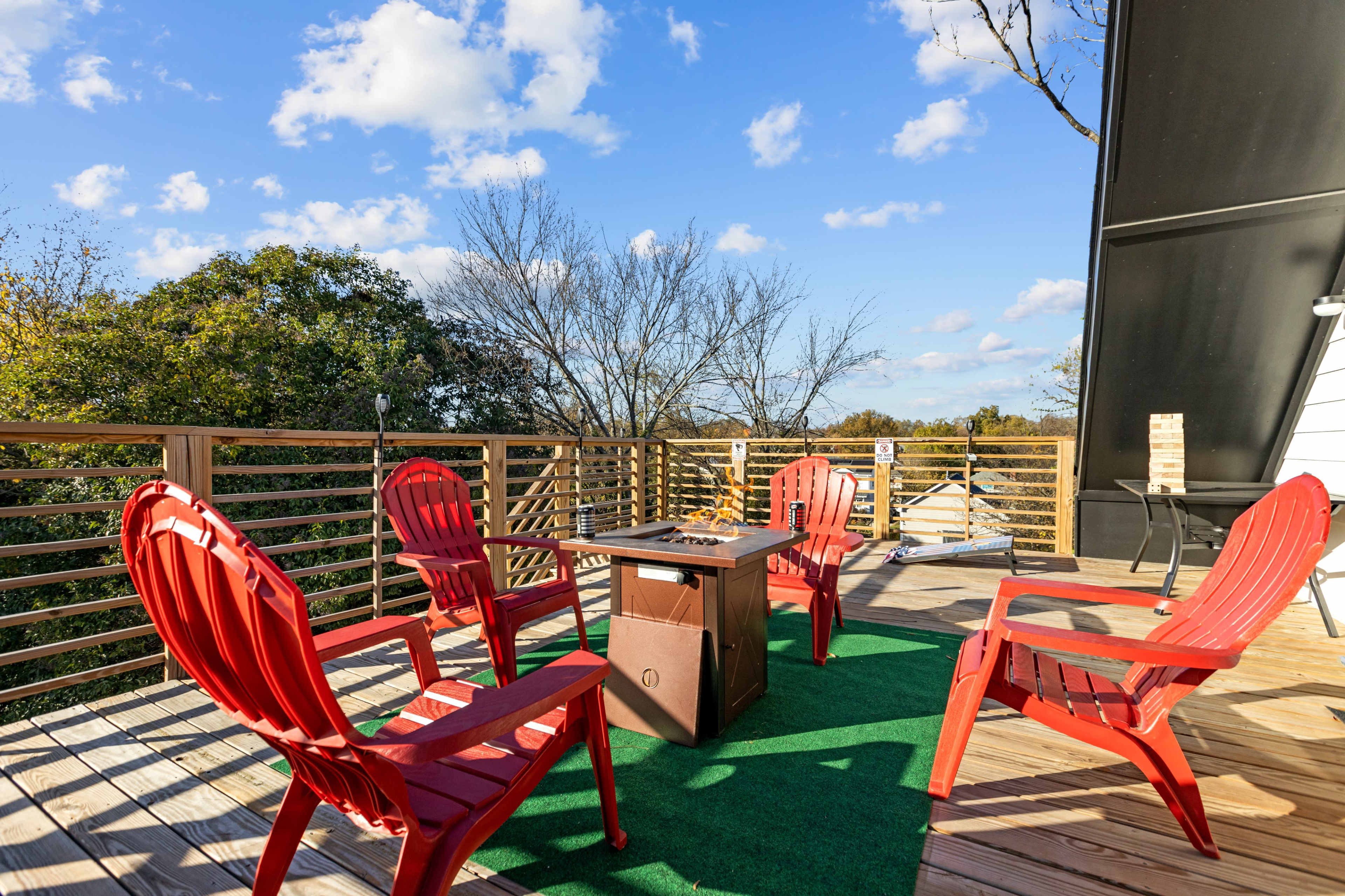 A deck features four red chairs arranged around a central table, with trees and a blue sky in the background.