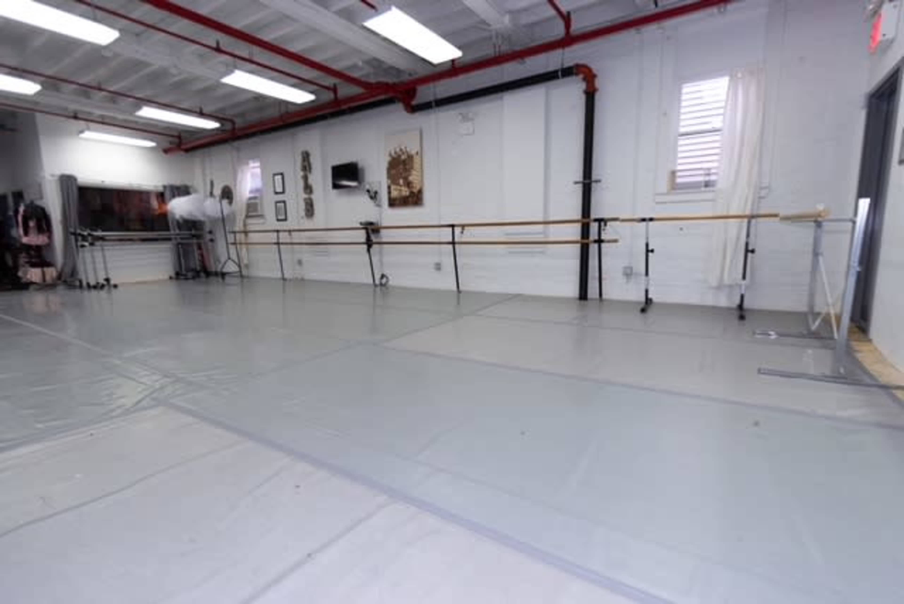 An empty dance studio with ballet bars along the walls and a grey floor covering.