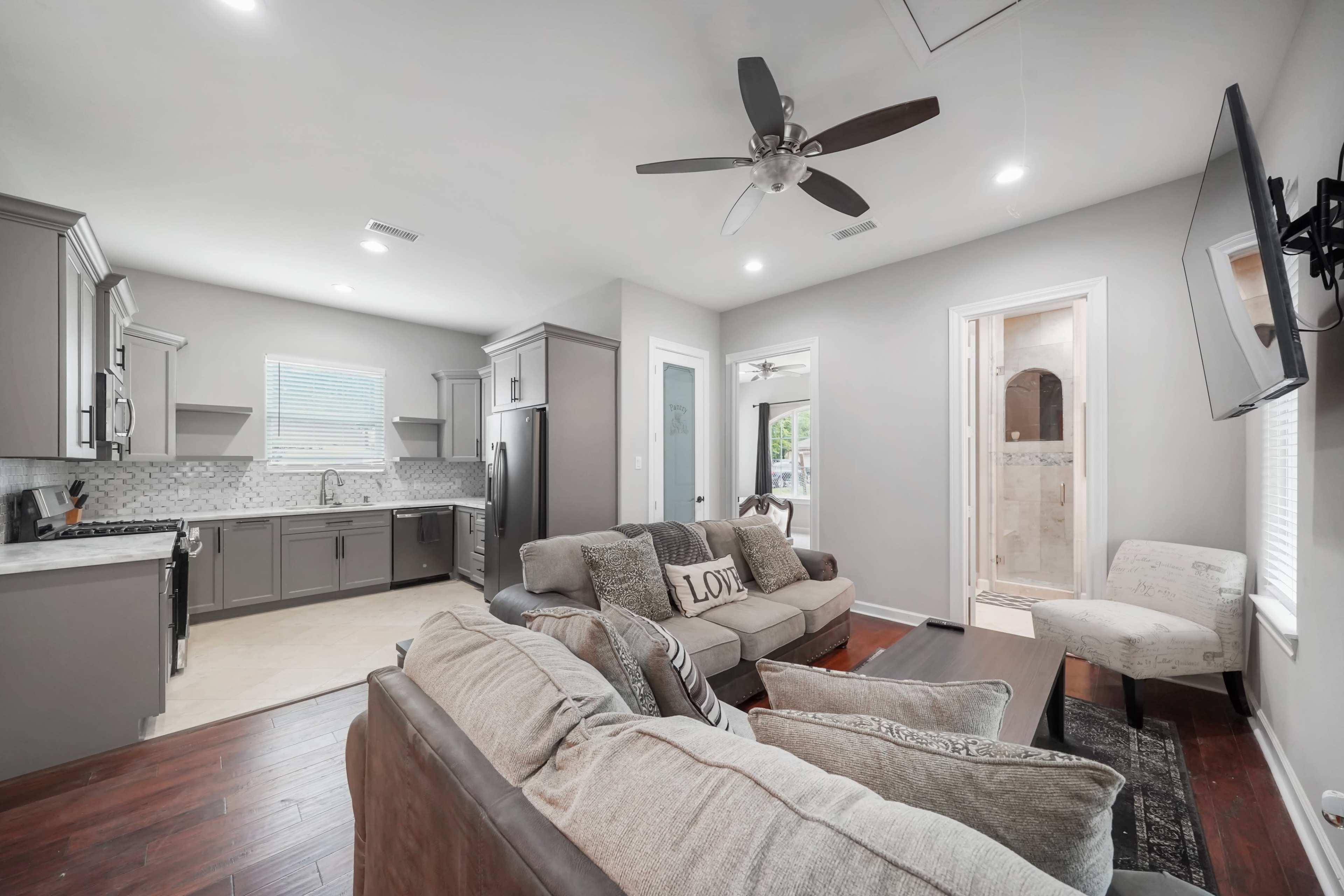A modern living room with a sofa facing a wall-mounted television, adjacent to a kitchen with gray cabinets and a dining area.