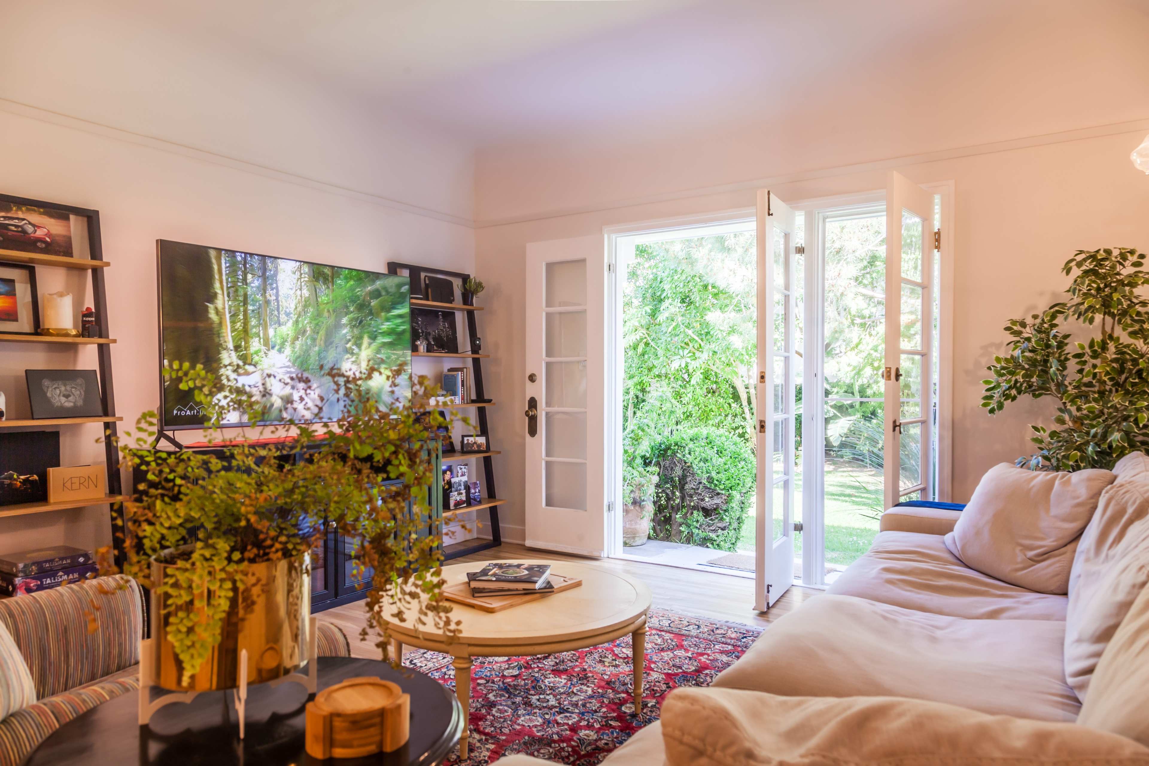 A cozy living room features a large television, a round coffee table, and a door open to a lush garden.