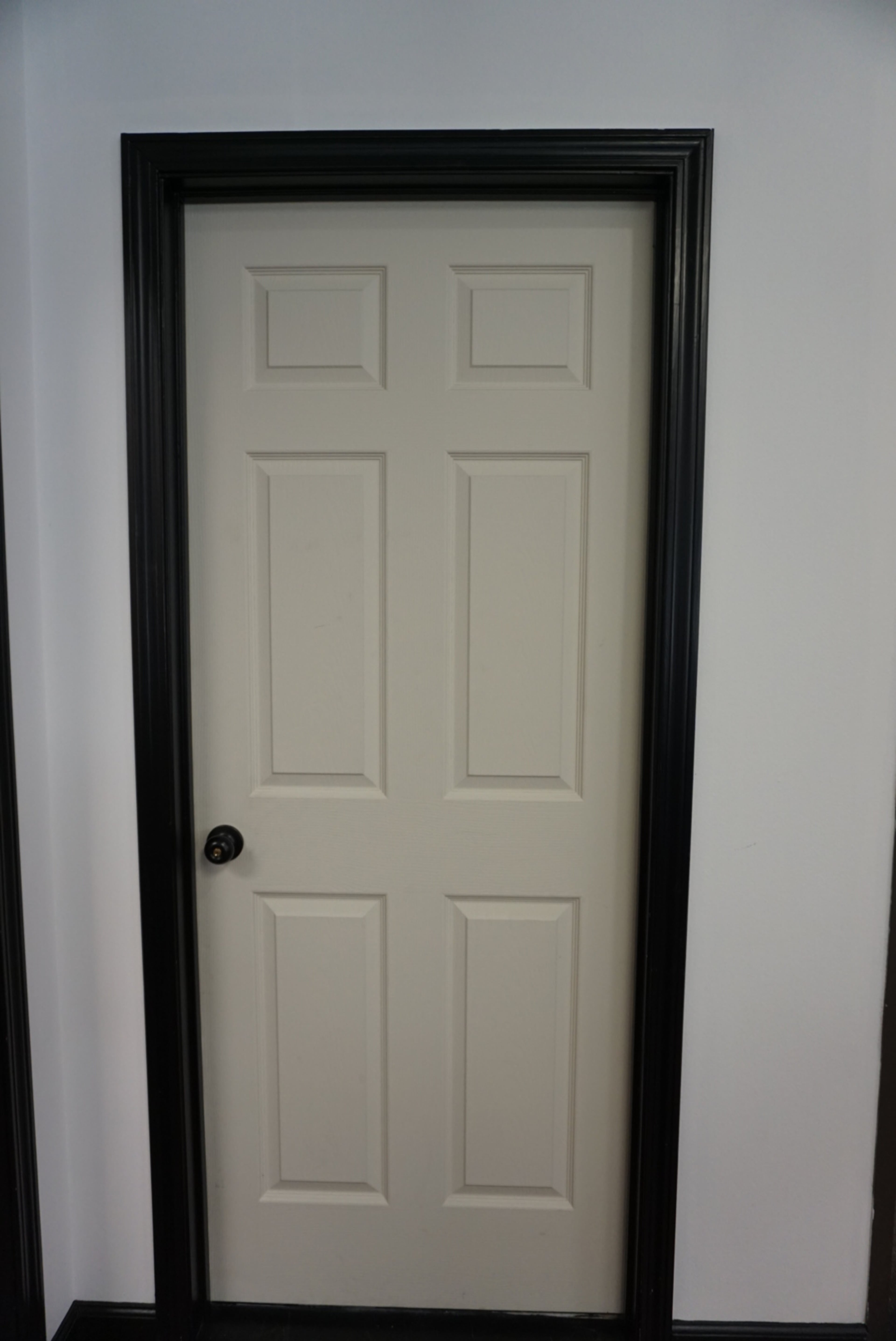 A closed six-panel door with a black doorknob, framed by black trim against a white wall.