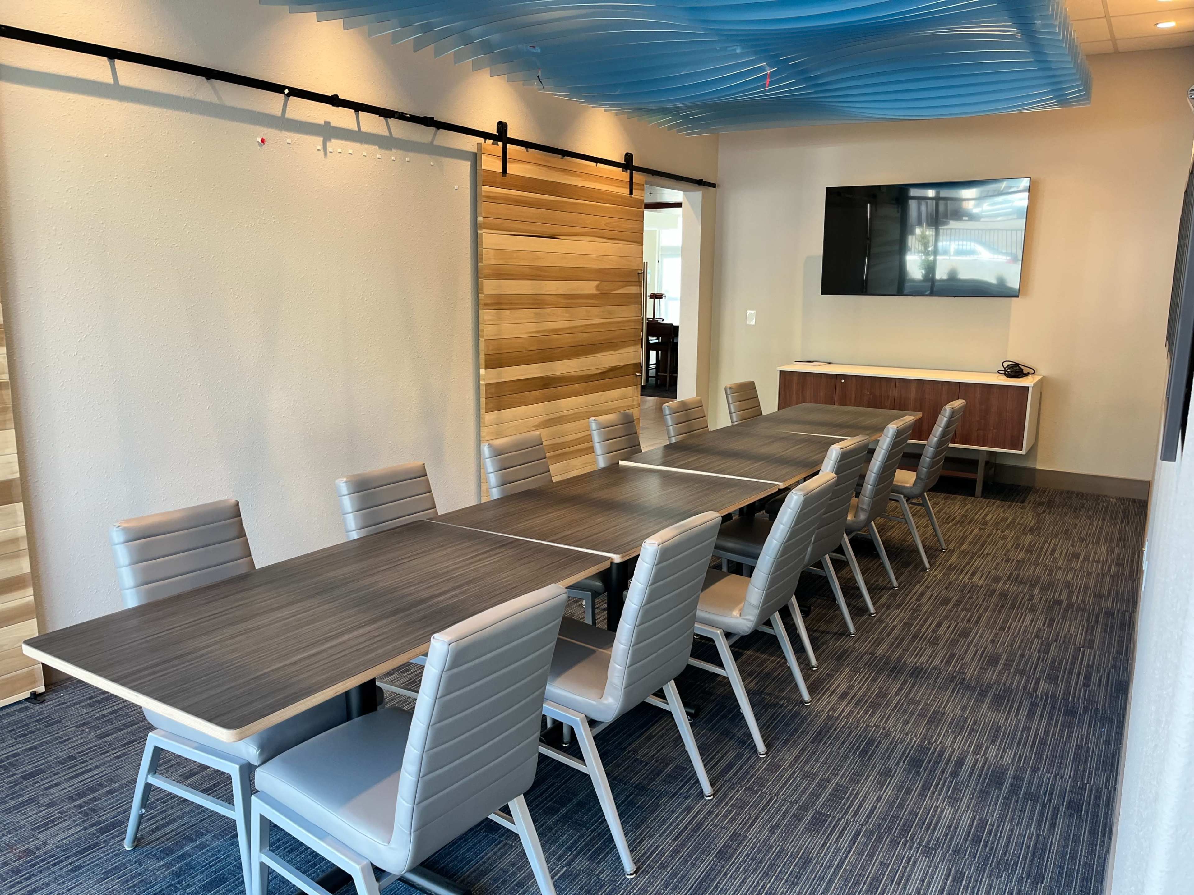 The image shows a modern conference room with two long tables arranged in a U-shape and several gray chairs, along with a wall-mounted television and a wooden accent wall.