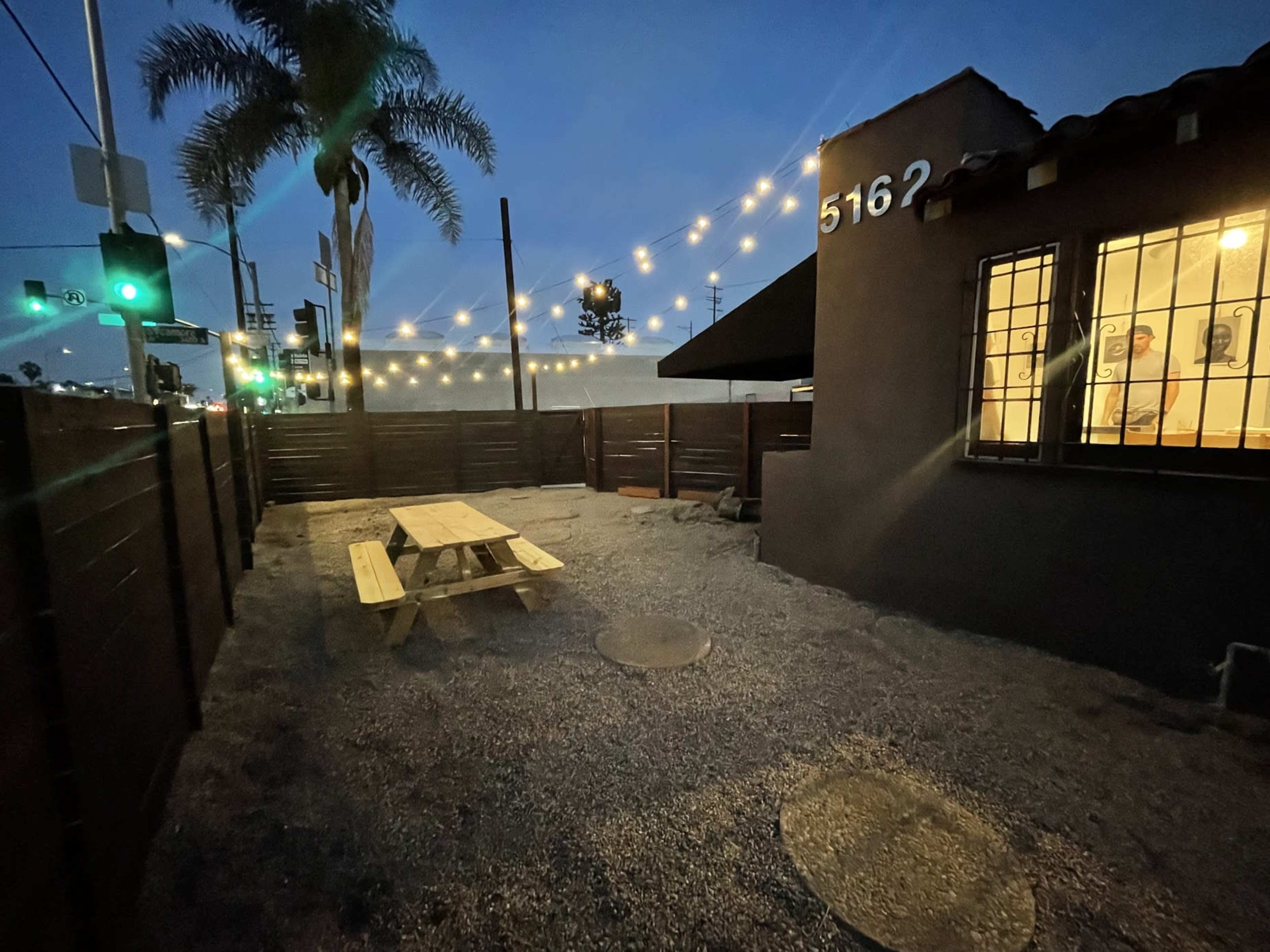 A quiet outdoor area with a wooden picnic table surrounded by gravel, illuminated by string lights against a dusky sky.