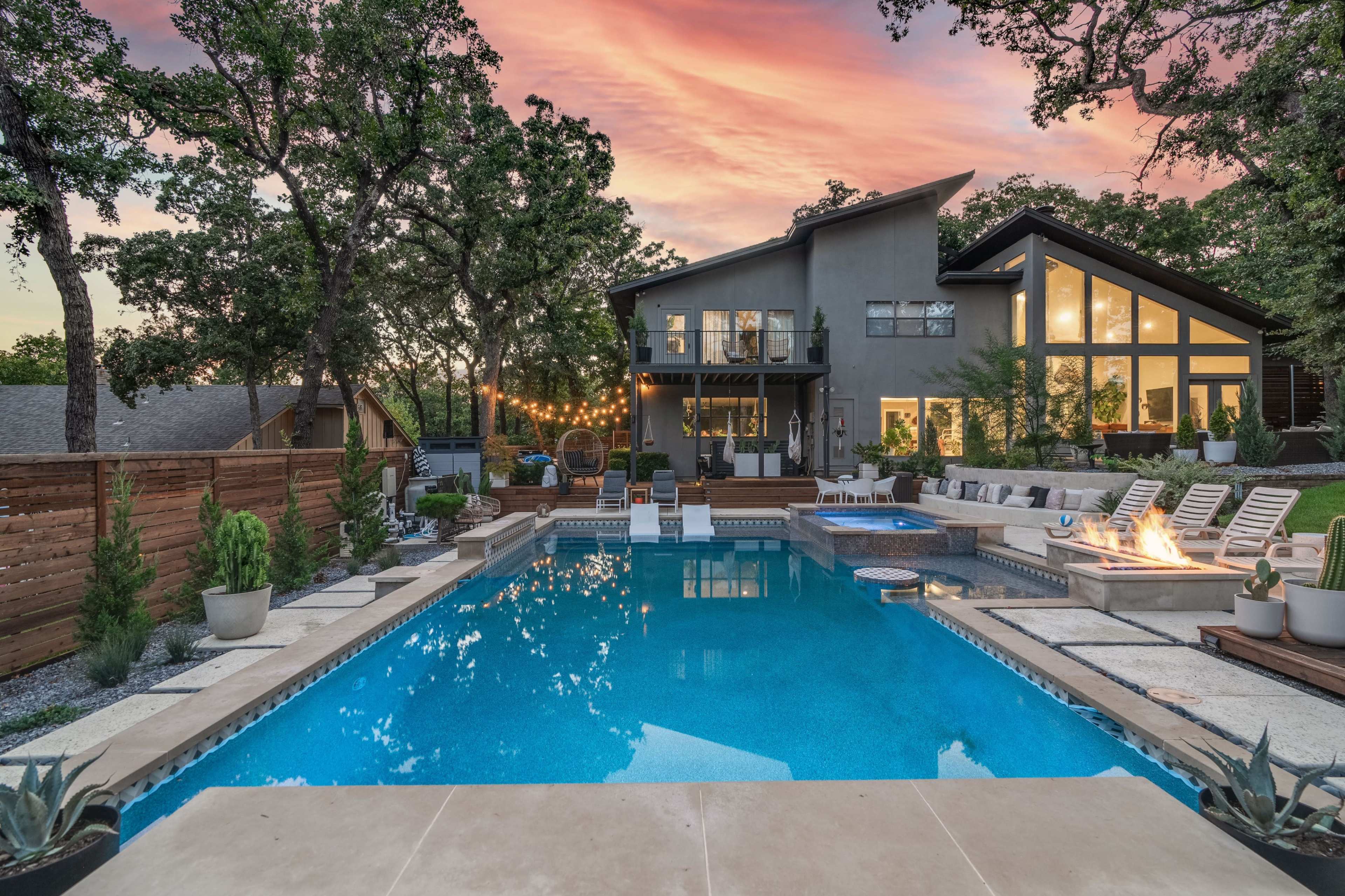 A modern home with large windows overlooks a blue swimming pool surrounded by lounge chairs, outdoor lighting, and landscaped greenery at sunset.