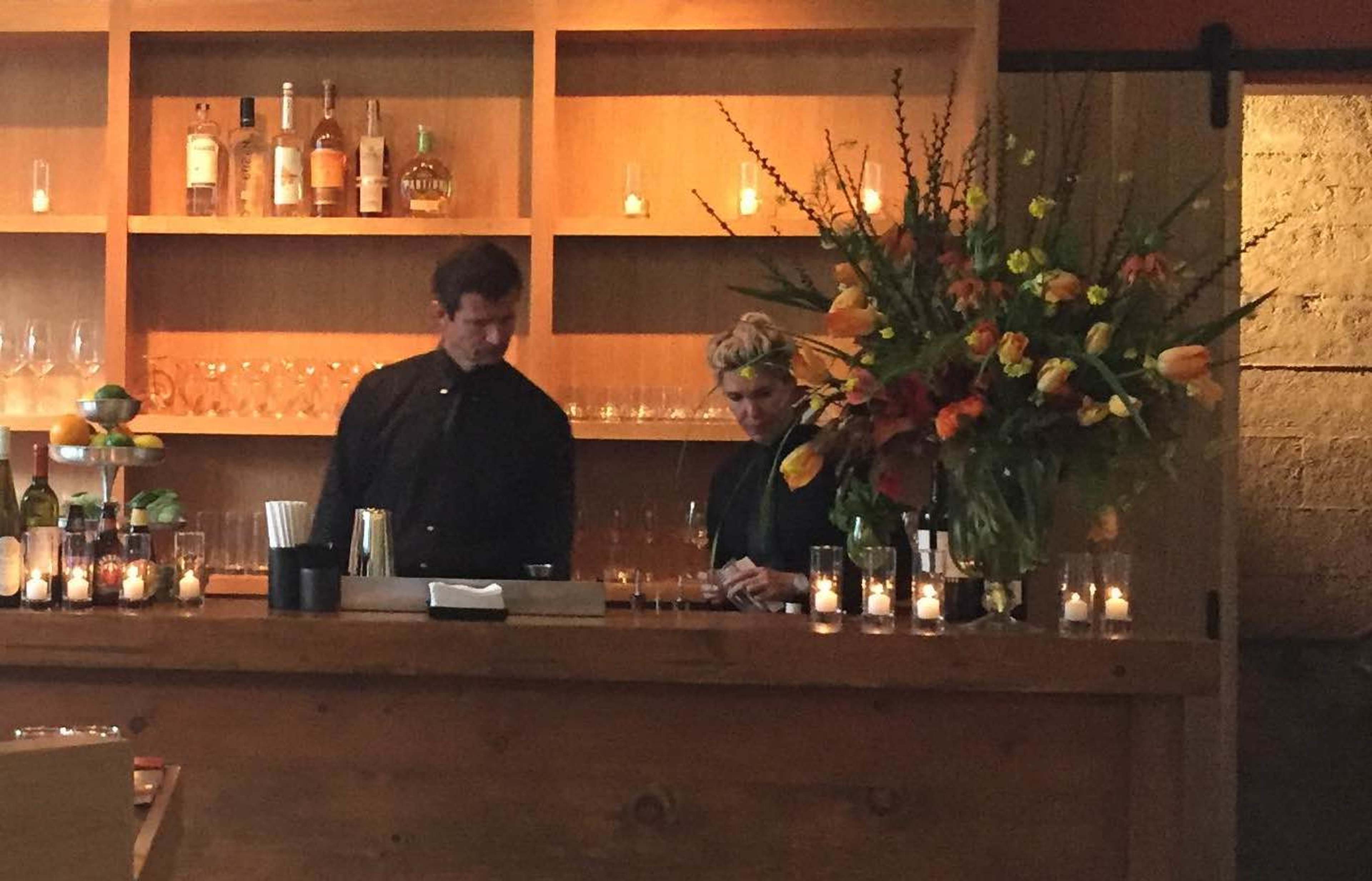 Two bartenders stand behind a wooden bar, where various bottles and fresh flowers are displayed, along with lit candles creating a warm ambiance.