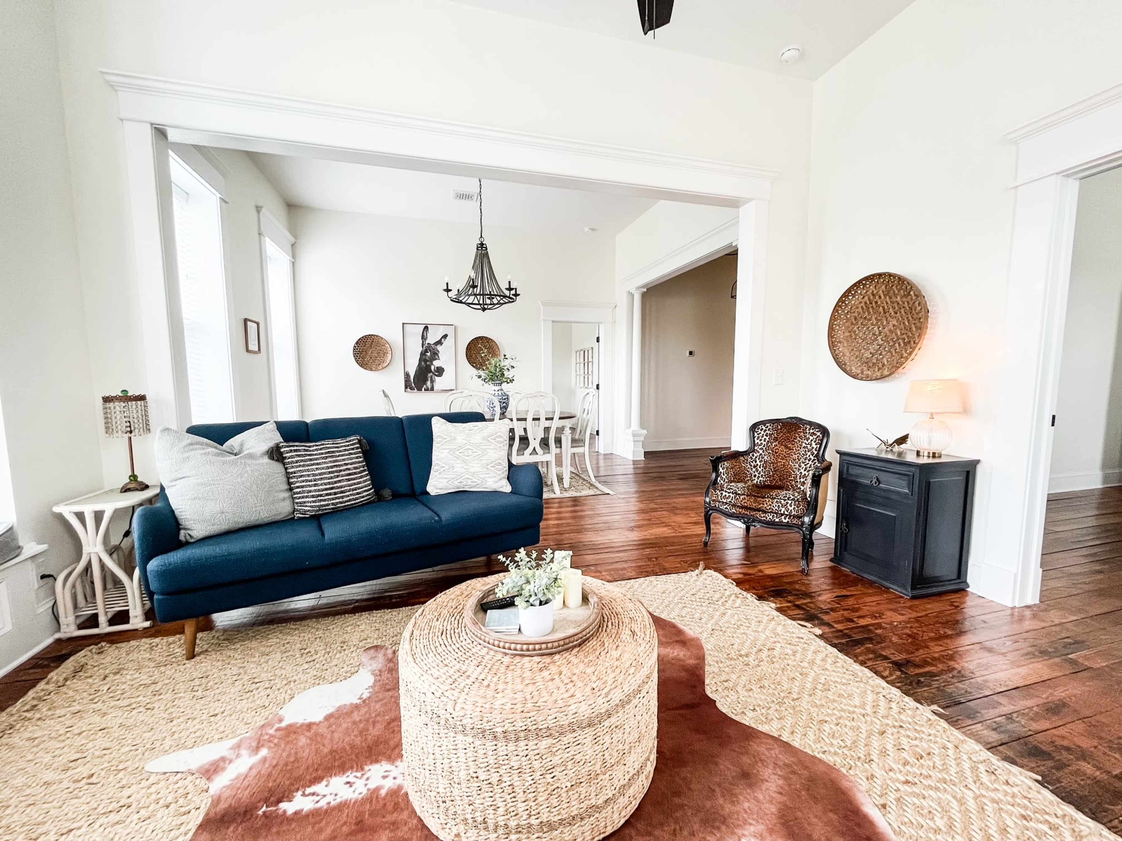 A living room features a blue sofa, a round woven coffee table, and a traditional chair, all set against wooden floors and light-colored walls.