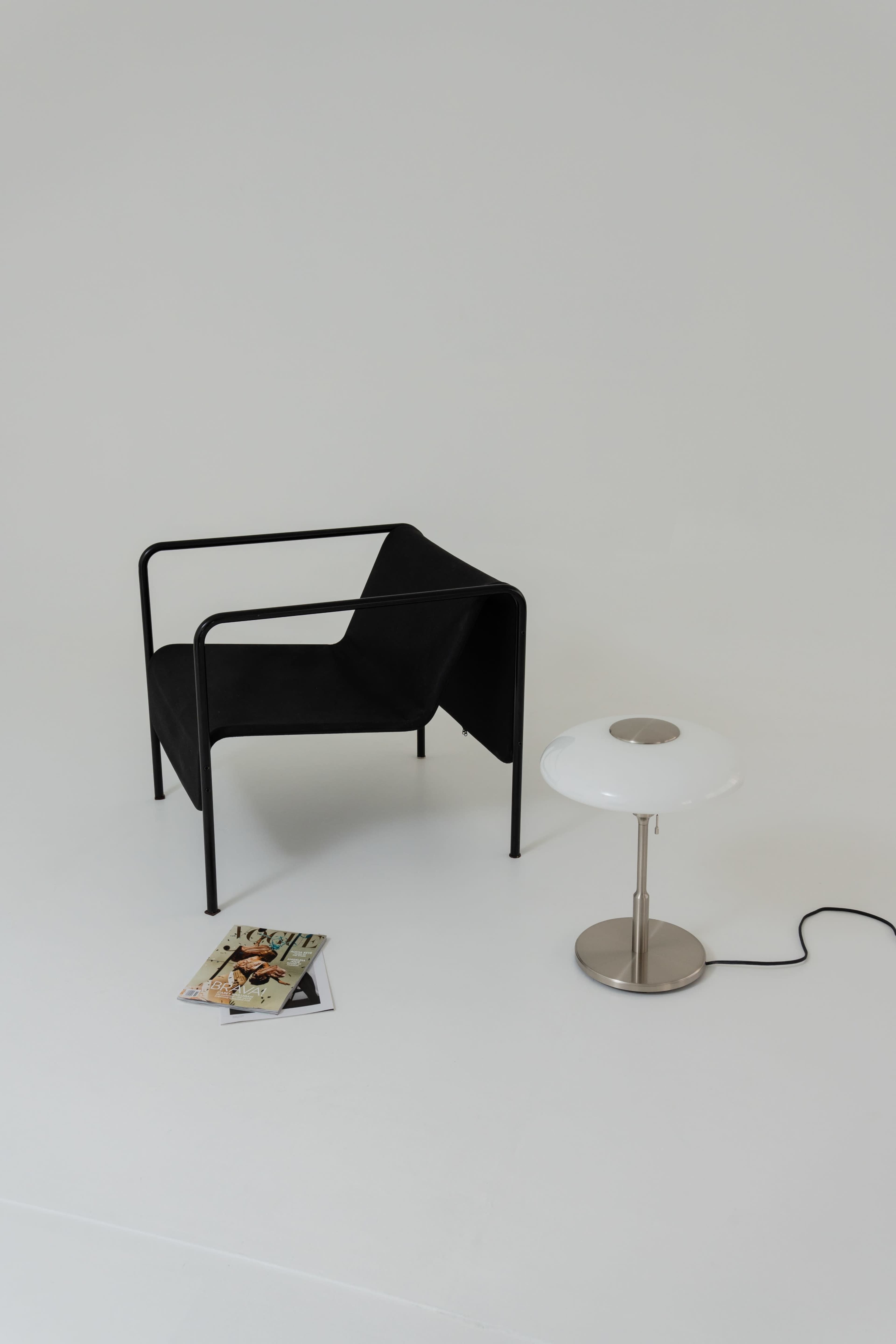 A black chair with a minimalistic design sits next to a white lamp on a light-colored floor, accompanied by scattered magazines.