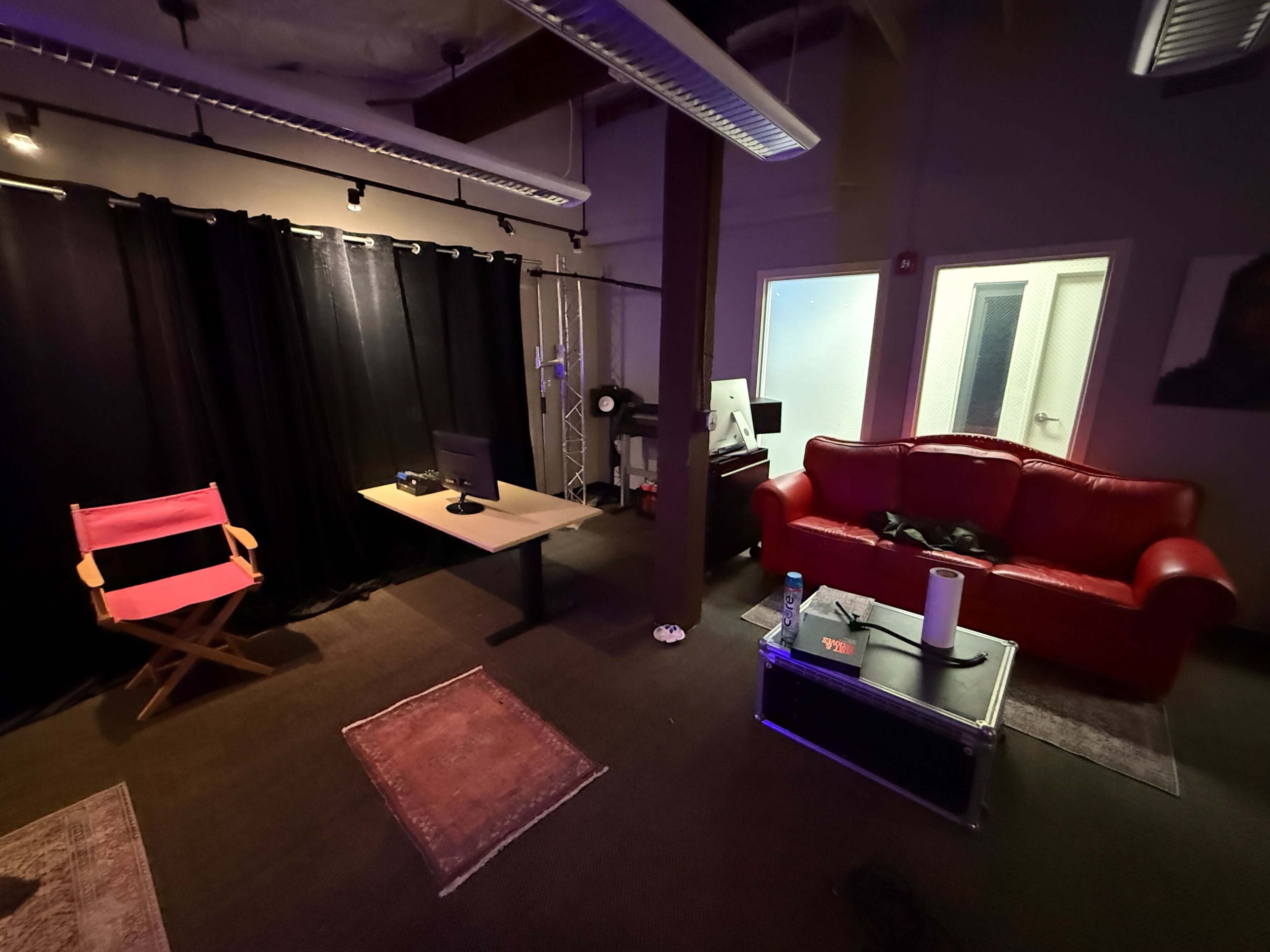 The image depicts a dimly lit room featuring a red couch, a desk with a computer, and a director's chair, with dark curtains and a couple of glass doors visible in the background.