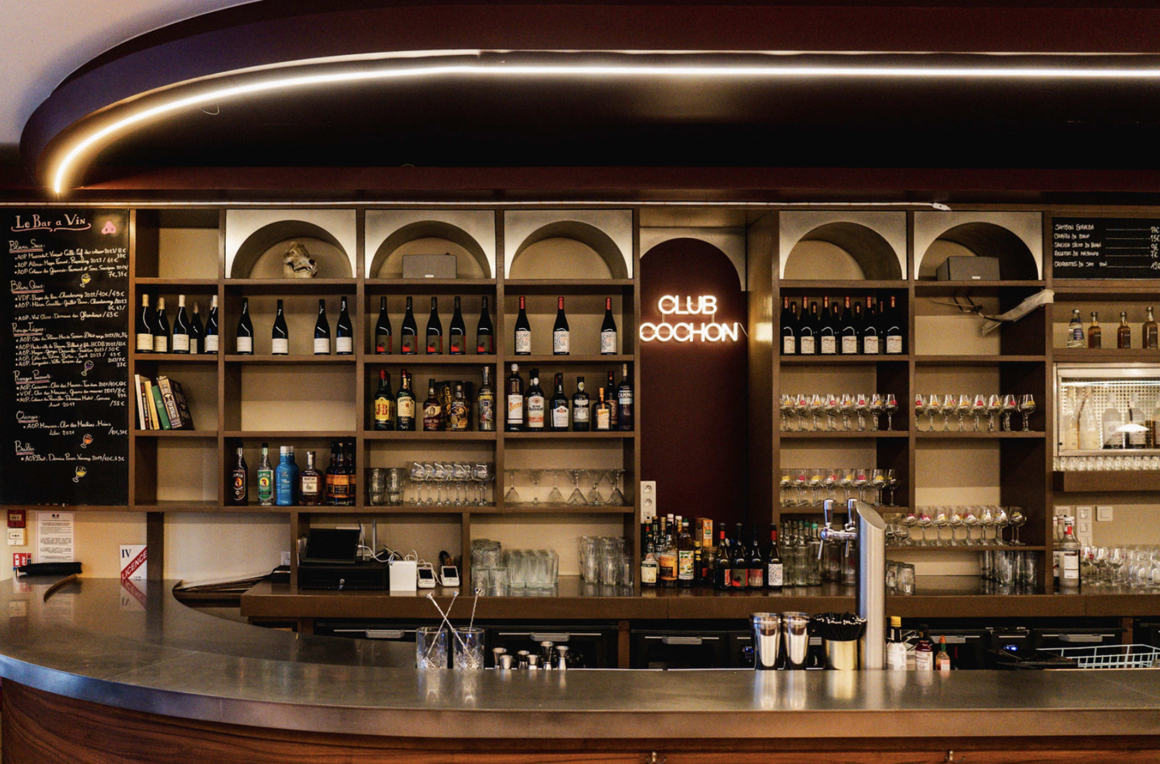 The image shows a modern bar setup featuring shelves stocked with various bottles, glasses, and a neon sign that reads "Club Cochon."